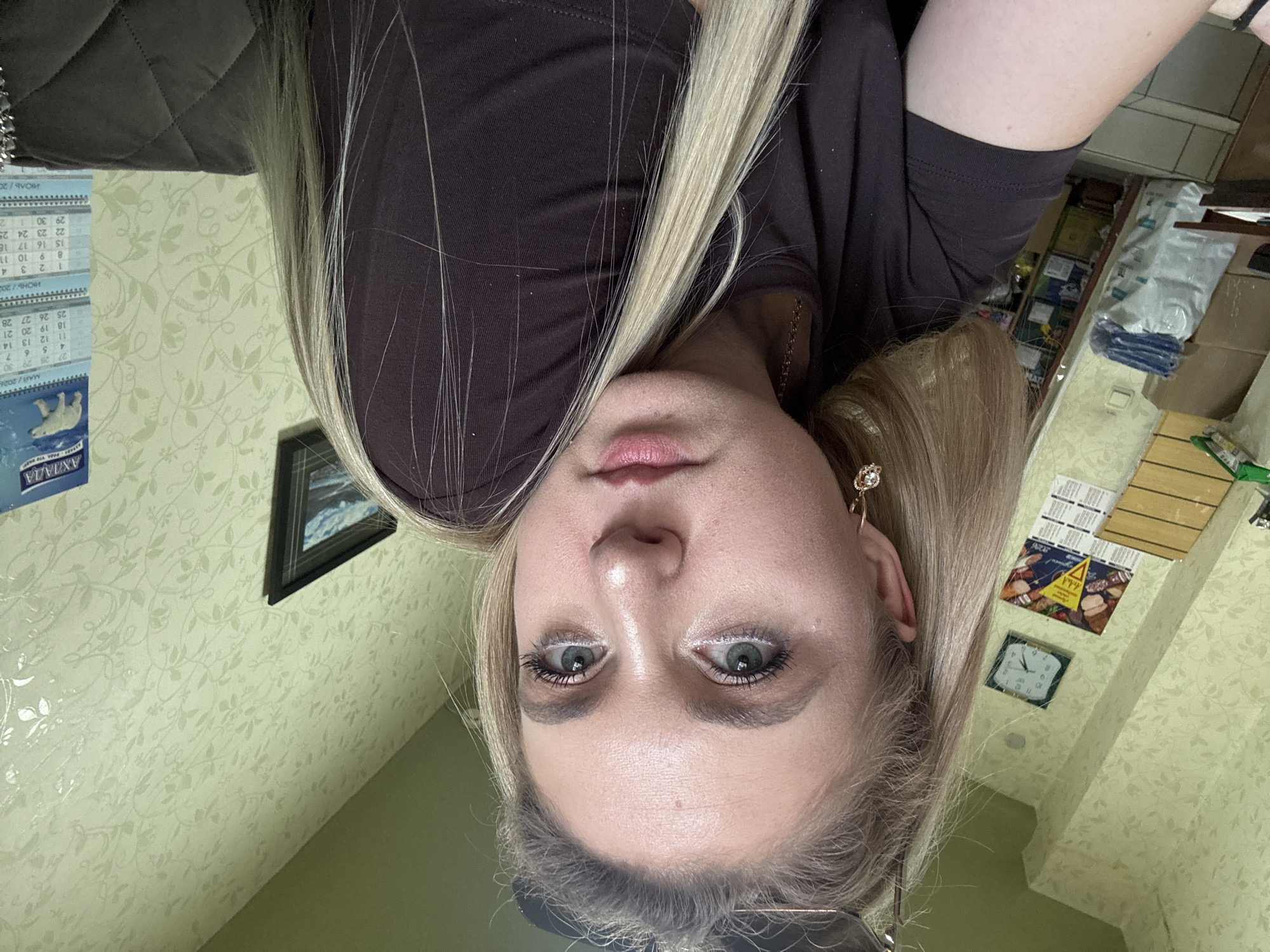 This is a vertical, close-up, point-of-view photograph of a young woman's face, taken from a low angle. She has blonde hair and is looking directly at the camera. Her eyes are open, and she has noticeable eye makeup, including white eyeliner and mascara. Her lips are slightly parted and appear to be glossed. She is wearing a dark brown top, and a glimpse of a quilted brown jacket is visible on the left side of the frame. A gold earring with a gem is visible in her right ear.
The background is out of focus but appears to be an indoor setting. To the left, a calendar is partially visible with Russian text and dates, indicating months such as "МАЙ/2026" (May/2026) and "ИЮНЬ/200" (June/200). There's also a picture of a polar bear on the calendar. On the right, shelves with various items are visible, including boxes and what appear to be food packages. A small square clock with Roman numerals is mounted on the wall. The wallpaper has a light green, floral pattern. The overall lighting suggests it could be daytime, but without clear windows or outdoor views, the time of day is not definitively ascertainable. The woman's expression is neutral.