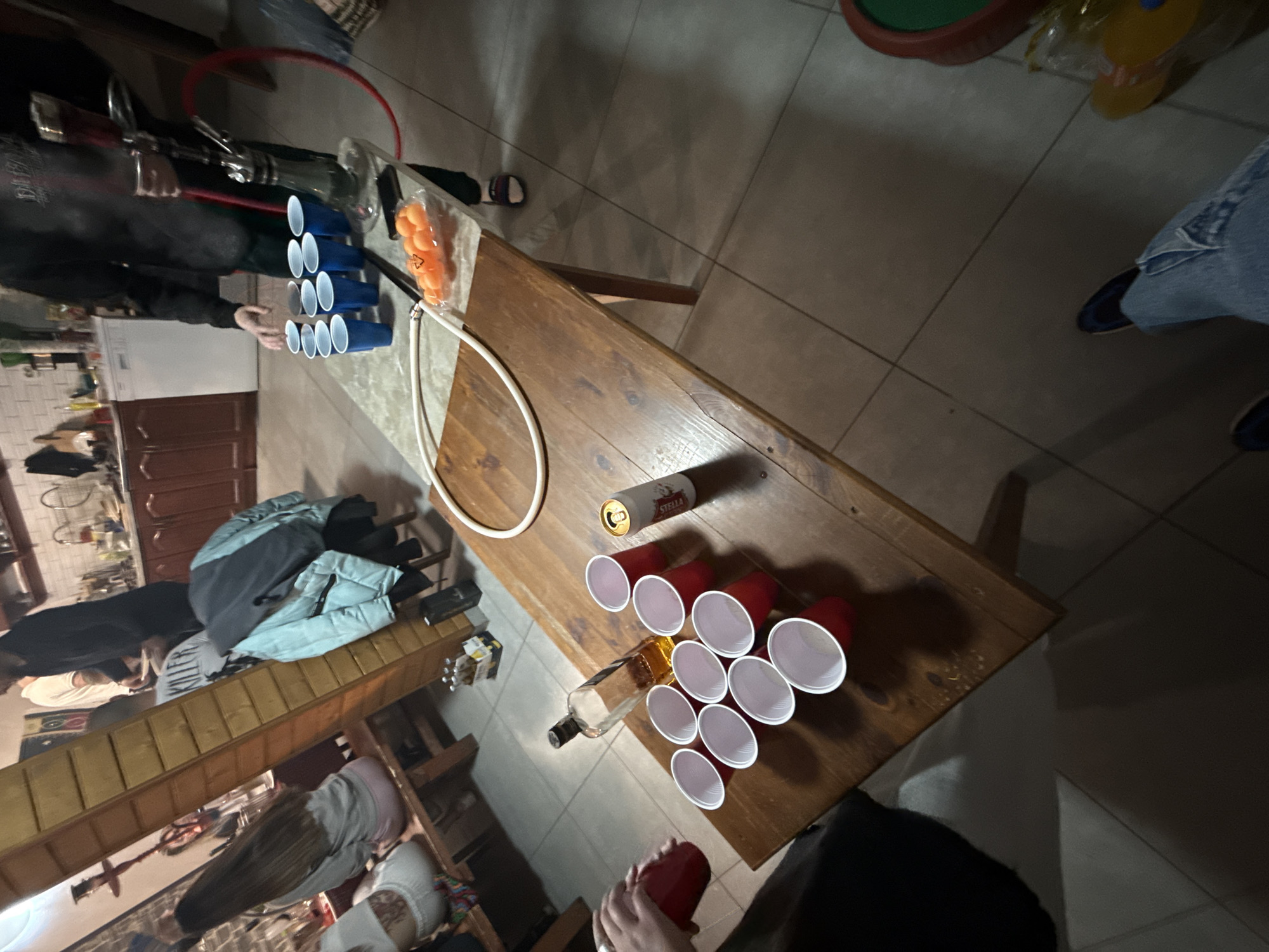 The image shows a scene of a beer pong game in progress. A wooden table is set up with two triangles of cups at opposite ends; one set is blue and the other is red. Ping pong balls are scattered near the blue cups. There is a beer can and a liquor bottle on the table. People are standing around the table, participating in the game. The background suggests an indoor setting, possibly a home, with a hula hoop resting on the table. The image does not provide information about the location in the city of Ribaritsa, Bulgaria.