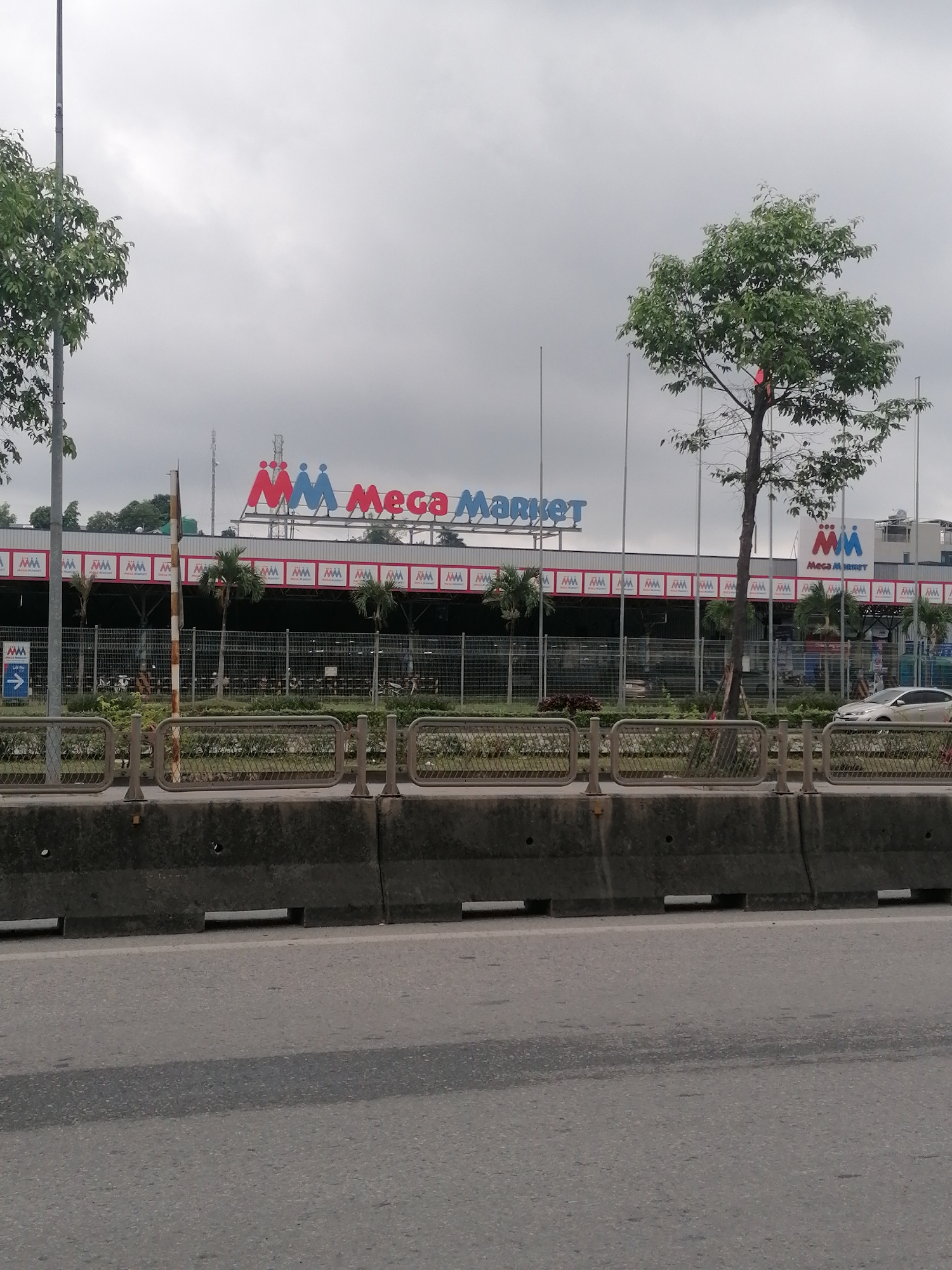 The image displays the exterior of a "Mega Market" store under an overcast sky, observed from a roadside perspective, likely in Ha Tu Ward, Vietnam. In the foreground, a grey asphalt road leads to a dark concrete barrier with rectangular cutouts. Beyond this barrier, a metal fence separates the roadway from a landscaped area featuring green bushes and several palm trees.
The main subject is the large commercial building of the Mega Market, adorned with a prominent red and blue sign on its rooftop displaying the text "Mega Market" alongside a stylized "MM" logo resembling two people. Below this, the building's facade features a repeating pattern of smaller "MM" logos and "Mega Market" text on a red and white striped banner. Another white box sign with the "MM" logo and "Mega Market" text is visible on the far right side of the building. A smaller blue directional sign with an arrow and "Mega Market" text is attached to the fence on the left.
Trees with green foliage are present on both sides of the frame, reaching towards the cloudy sky, with one tall tree on the far left and another on the right, partially obscuring the market building. A white car is partially visible near the market's entrance on the right, and indistinct shapes suggesting parked motorbikes can be seen through the fence in the market's parking area. The weather is cloudy, and the overall scene depicts a functional commercial establishment on a typical day, with no specific human activity or event taking place.