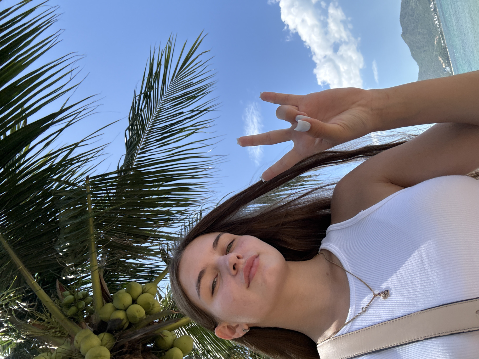 A young woman with long brown hair, wearing a white tank top and a gold necklace with a small pendant, takes a selfie outdoors. She is looking at the camera with a neutral expression and has her right hand raised, forming a rock-on gesture with her index and pinky fingers extended. Her fingernails are painted white. Behind her, a palm tree with green coconuts is visible, as is a bright blue sky with scattered white clouds. In the background, there is a body of water, a shoreline with buildings, and green hills. The scene suggests a tropical location, likely a beach in Nha Trang, Vietnam, given the palm trees, ocean, and hills characteristic of the region.