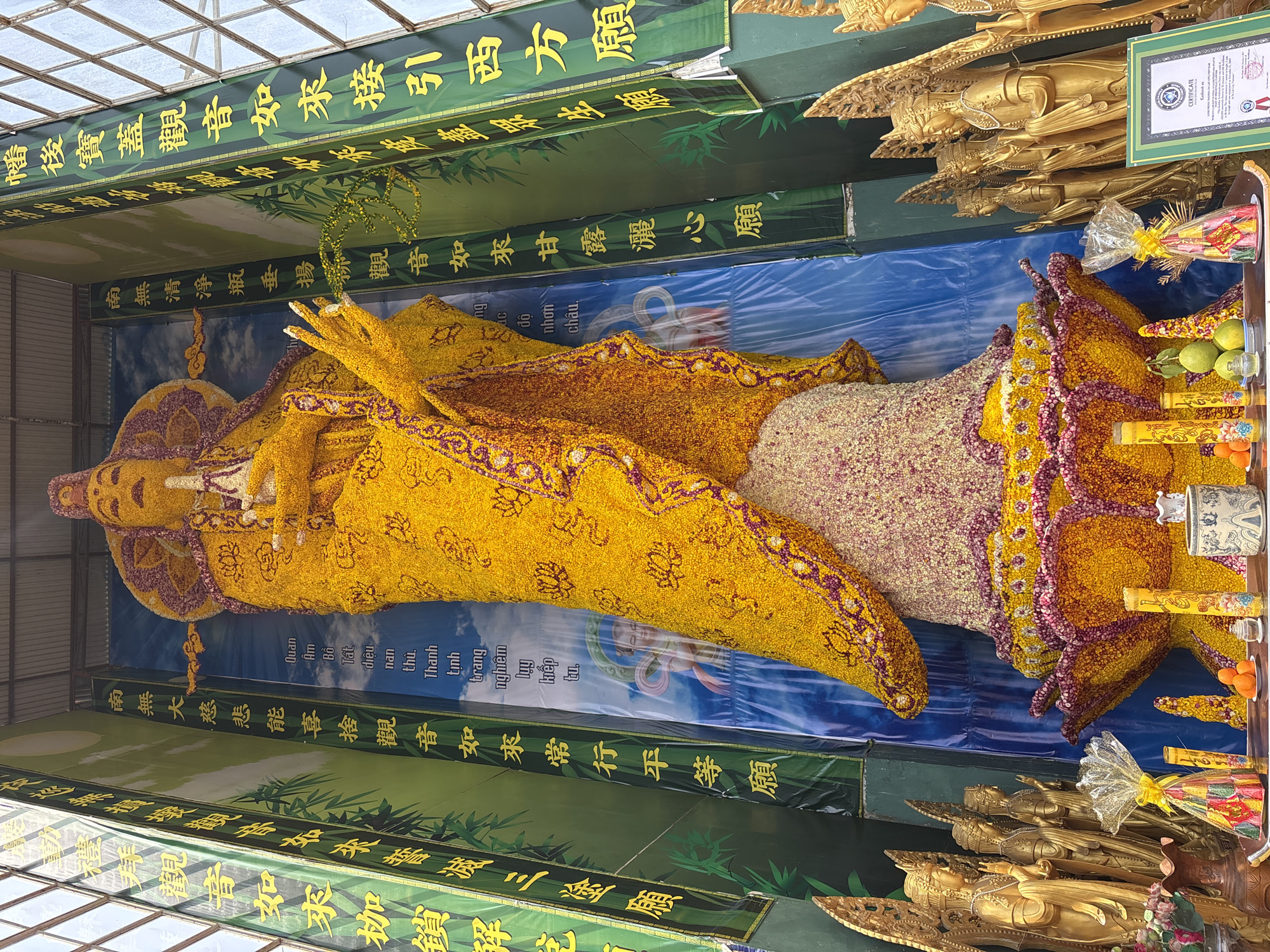 The image shows a large reclining figure of a deity, possibly Buddha or Quan Yin, constructed entirely of yellow and other-colored flowers. The figure lies on a blue background. Banners with Chinese/Vietnamese characters hang above and below the figure. In the foreground, there are gold statues. A certificate or document is displayed above the head of the figure. The text includes "CERTIFICATE" and the city "Xuân Thành," suggesting the image was taken there. The scene is likely indoors, possibly in a temple or exhibition space.