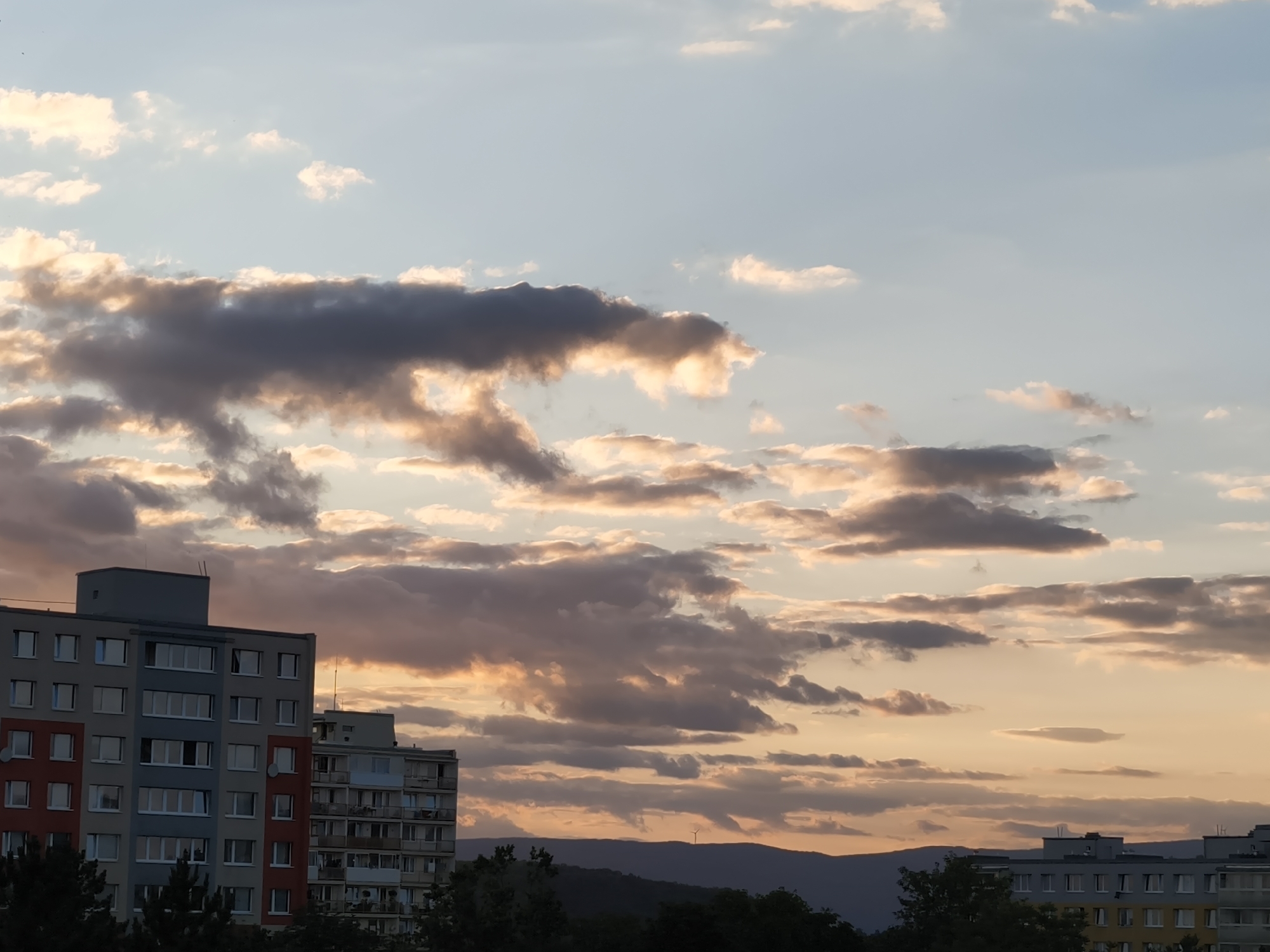 The image depicts a sunset over the city of Most, Czechia. In the foreground, two residential apartment buildings are visible. The building on the left is taller and features a multi-colored facade with sections of red, grey, and white. The building on the right appears slightly shorter and has a more uniform facade with balconies. Below the buildings, the tops of trees are visible, forming a dark green band across the bottom of the frame. In the background, rolling hills are silhouetted against the vibrant sky. The sky is filled with dramatic clouds, ranging in color from pale blue and white to shades of grey and orange, indicating the setting sun. A small wind turbine is visible on one of the distant hills. The scene is serene and captures the transitional light of dusk.