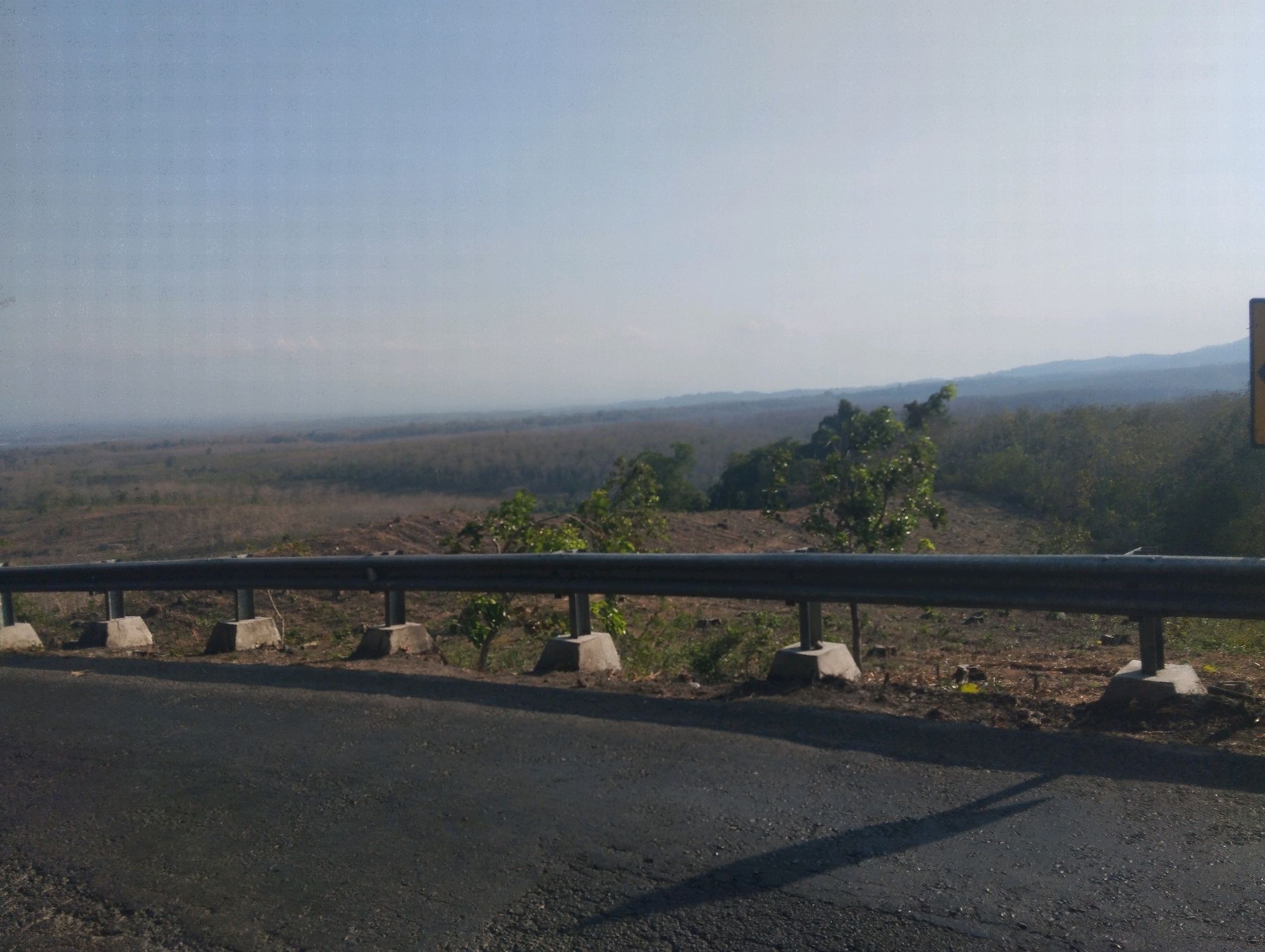 The image shows a roadside view overlooking a vast, dry landscape under a hazy, pale blue sky. A metal guardrail runs horizontally across the foreground, supported by concrete posts. The road surface in front of the guardrail appears to be asphalt, with some texture and debris visible.
Beyond the guardrail, the scene unfolds as a wide expanse of dry, brown vegetation, interspersed with patches of darker green foliage. The land appears to be somewhat cleared in places, suggesting potential agricultural use or recent deforestation. In the distance, the landscape softens into a muted, hazy horizon with rolling hills or mountains visible. The overall impression is of a rural or undeveloped area, possibly in a tropical or subtropical region, given the dry appearance and vegetation.
The lighting suggests it is daytime, likely late morning or afternoon, with the sun casting shadows on the road. The haze in the sky could indicate a dry season, dust, or pollution. There is a yellow traffic sign with a black arrow visible on the right side of the image, providing a subtle clue to the location being along a road with a bend. No people are visible in the image. The setting feels expansive and somewhat desolate due to the dryness and the sheer scale of the landscape.