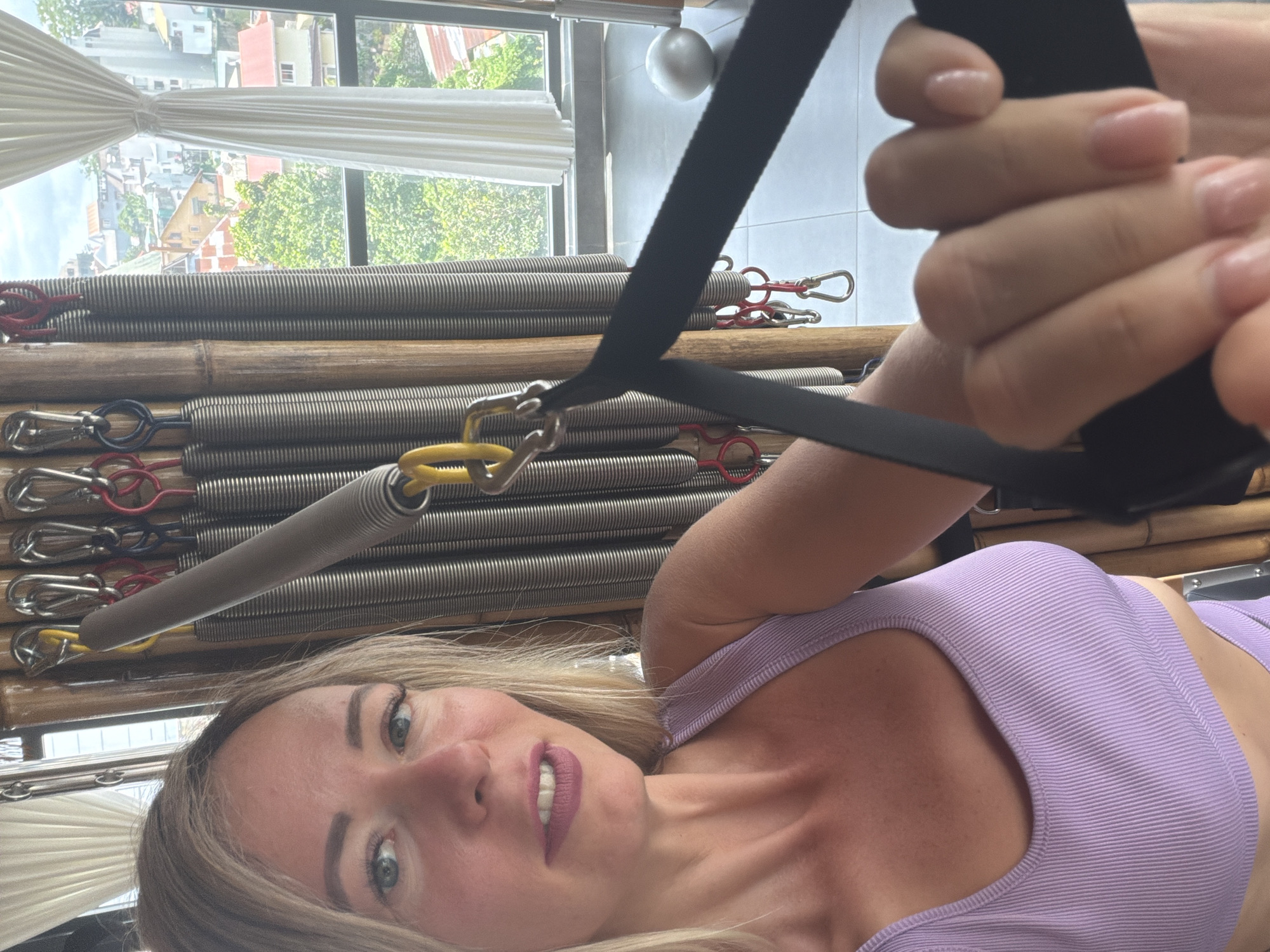 A woman with blonde hair and blue eyes, wearing a light purple sports bra, is holding a black strap attached to a pilates reformer. She is taking a selfie in what appears to be a studio with large windows overlooking a city. The windows reveal buildings with red and yellow roofs, lush green trees, and white curtains. A small white ball rests on the floor near the window. The pilates reformer has wooden posts and several metal springs of varying colors (red, blue, yellow) attached by carabiners. The background shows the city of Da Lat, Vietnam, with its distinctive architecture and hilly landscape.