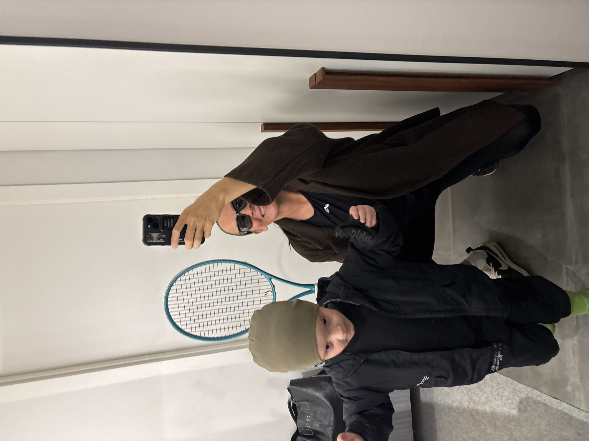 A woman and a child are visible in the image. The woman is holding a smartphone up, presumably taking a selfie. She is wearing sunglasses, a black top, and a brown coat. The child is wearing a beanie and a black jacket. A tennis racket is visible in the background. A black bag is at the bottom of the image. The image appears to be taken indoors, possibly in a fitting room or hallway. The exact location and connection to Chaikovsky, Russia are not discernible from the image.