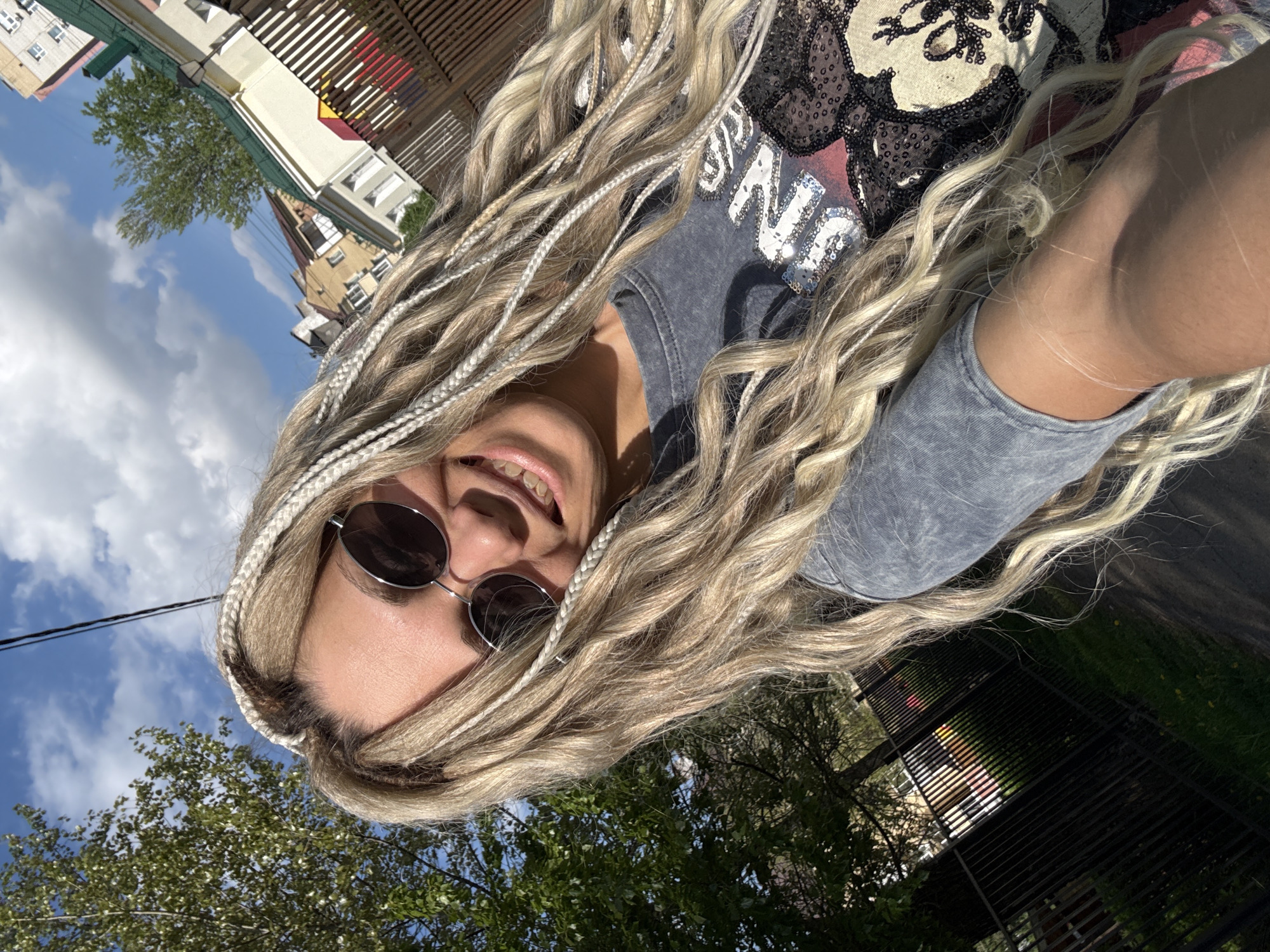 A medium shot depicts a woman with long, blonde hair, some of which is styled into braids, wearing round sunglasses and a gray t-shirt, smiling at the camera. The background includes buildings, trees, and a blue sky with white clouds. The location appears to be outdoors, likely a street or park in a city. The presence of buildings and trees, along with the sunny weather, suggests an urban environment in the Заволжье region of Russia.