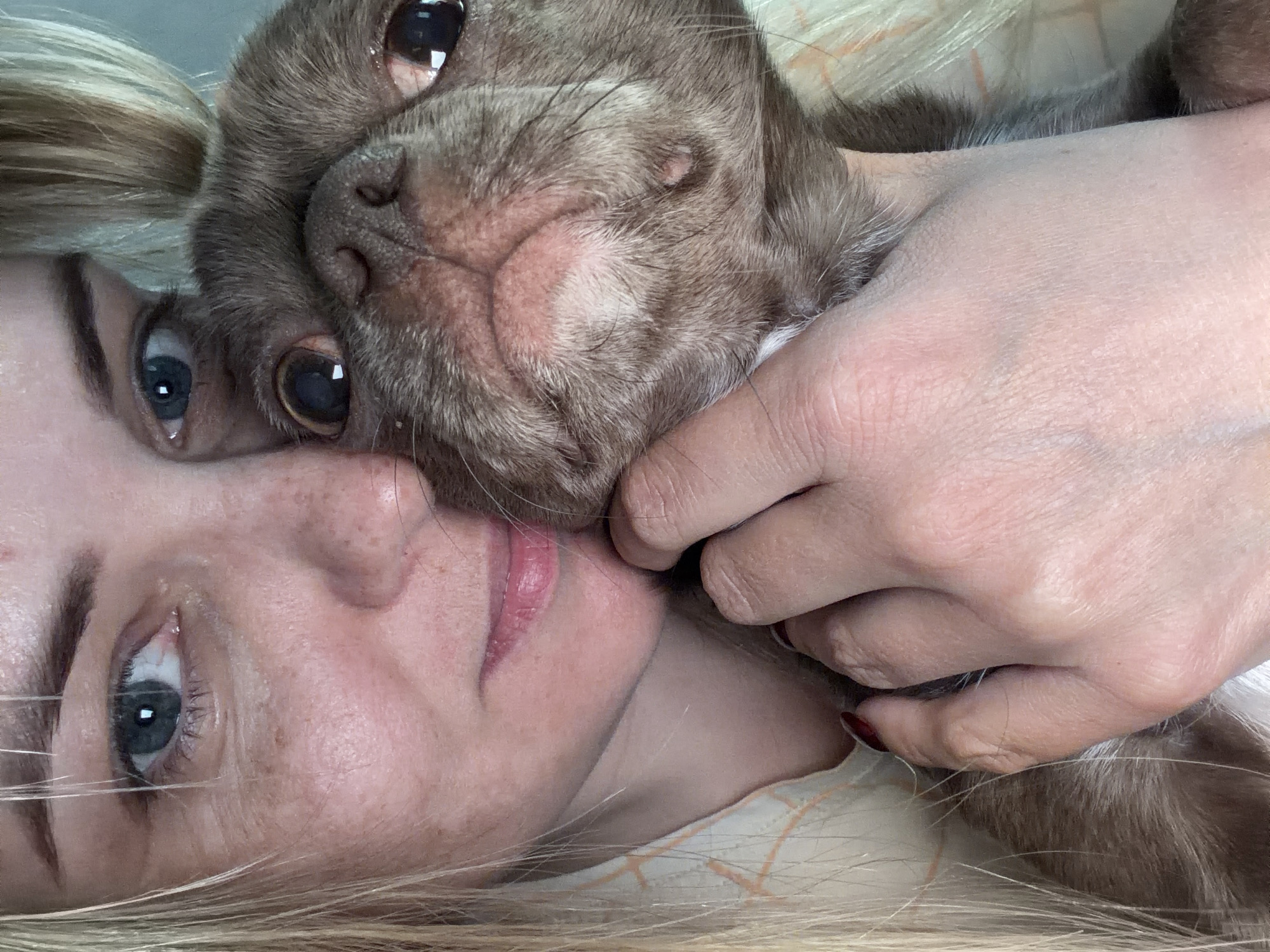 A woman with blonde hair and blue eyes lies on her back, face to face with a brown pug dog. Her right hand is around the dog's neck. The woman's face is tilted slightly towards the camera, with her nose near the dog's face. The dog's face is close to the camera, with its large eyes visible. The background is blurred and indistinct. There is no indication of Votkinsk, Russia in this image.