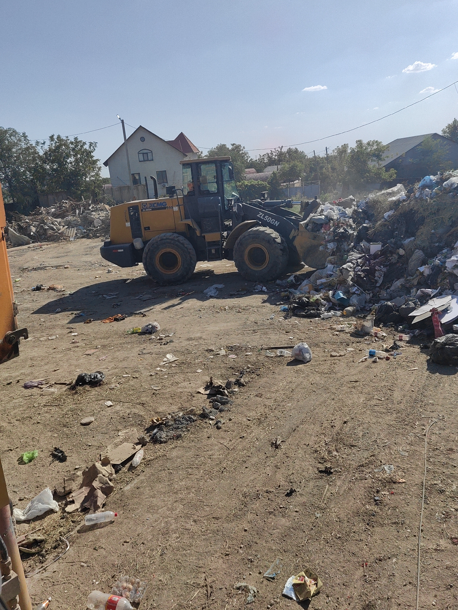 This image depicts an outdoor scene in the city of Shymkent, Kazakhstan, characterized by a significant amount of refuse. A large yellow wheel loader, identified by the "XCMG ZL50GN" marking, is positioned prominently, actively engaged in or near a large pile of trash. The loader has its bucket lowered, suggesting it is in the process of moving or collecting waste. Surrounding the loader and the refuse pile is a dirt ground area littered with various discarded items, including plastic bottles, bags, and debris. In the background, there are residential buildings and trees, indicating a potentially semi-urban or rural fringe location. The sky is clear and blue with scattered clouds, and the lighting suggests it is daytime. The overall impression is one of an active waste disposal or cleanup operation in a somewhat unkempt environment.