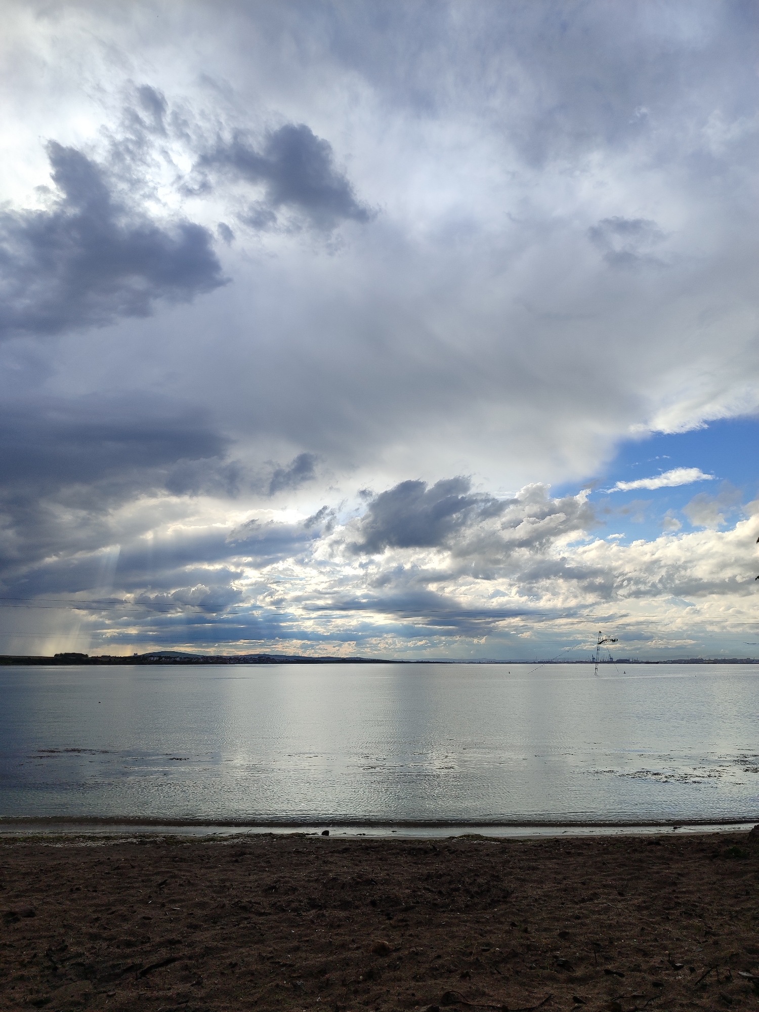 The image depicts a coastal scene, likely a beach, under a dramatic, cloudy sky. The foreground shows dark-colored sand, which appears to be wet. The middle ground is dominated by a body of water, likely the sea or a large bay, with a calm surface reflecting the sky. In the distance, a low-lying landmass with hints of buildings or structures is visible. The sky is filled with a mix of dark, stormy clouds and patches of brighter, partly cloudy areas. There are visible rays of light breaking through the clouds in the left side of the horizon. There are tower-like constructions in the background of the water body. The location is possibly in the city of Burgas, Bulgaria.