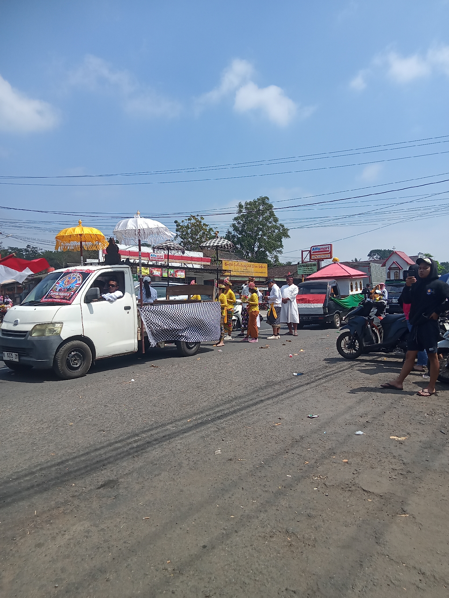 In Urutsewu, Indonesia, a procession moves along a paved road under a bright, partly cloudy sky. A white pickup truck, adorned with a checkered cloth on its bed and carrying decorative umbrellas, leads the procession. Two individuals are seated in the truck's cabin, one appearing to be driving. Several people in traditional attire, including women in yellow and orange clothing and men in white robes, walk beside and behind the truck. Motorcycles are parked on the side of the road, and a person in a black hoodie and shorts is visible in the foreground, looking at their phone. Buildings, including a red-roofed structure with a cross and a sign for "Alfamart," line the background. The road surface is asphalt, with some debris scattered on it.