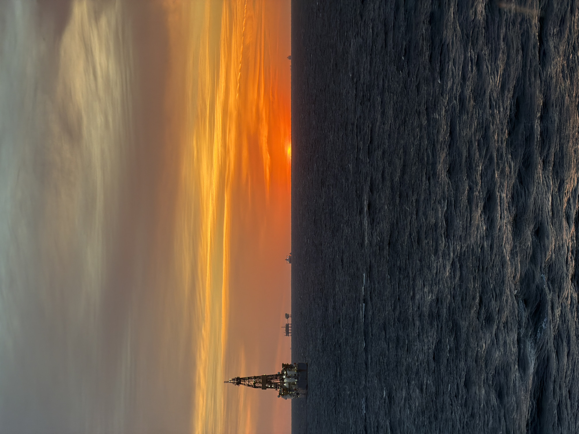 The image displays an expansive maritime scene at either sunrise or sunset, with a prominent horizon line dividing the sky from the ocean.
The sky, occupying the upper half of the frame, is bathed in the warm, intense hues of the rising or setting sun. A bright, orange-red sun is visible just above the horizon, casting a radiant glow. The colors in the sky transition from fiery orange and yellow near the horizon to softer, streaky clouds of grey, pale yellow, and hints of blue higher up. These long, wispy clouds create a dramatic effect across the expanse of the sky.
The lower half of the image is dominated by the vast, dark expanse of the ocean. The water appears deep blue-grey, almost black, with a textured surface indicating waves or ripples. Subtle reflections of the sky's light can be seen on the water's surface, particularly closer to the horizon.
Along the horizon, several industrial structures are silhouetted against the bright sky. In the foreground, on the left side, a large, complex offshore drilling rig or production platform is clearly visible. It features a tall derrick or mast structure and numerous lights illuminating its various decks and components, suggesting ongoing operations. Further in the distance, closer to the sun's position, at least three additional smaller, less distinct offshore platforms or vessels are visible as dark shapes against the brilliant backdrop.
The time of day is definitively either sunrise or sunset, as indicated by the low position of the sun and the vibrant colors in the sky. The scene captures the intersection of industrial activity and natural beauty in a marine environment. There is no visible text in the image.