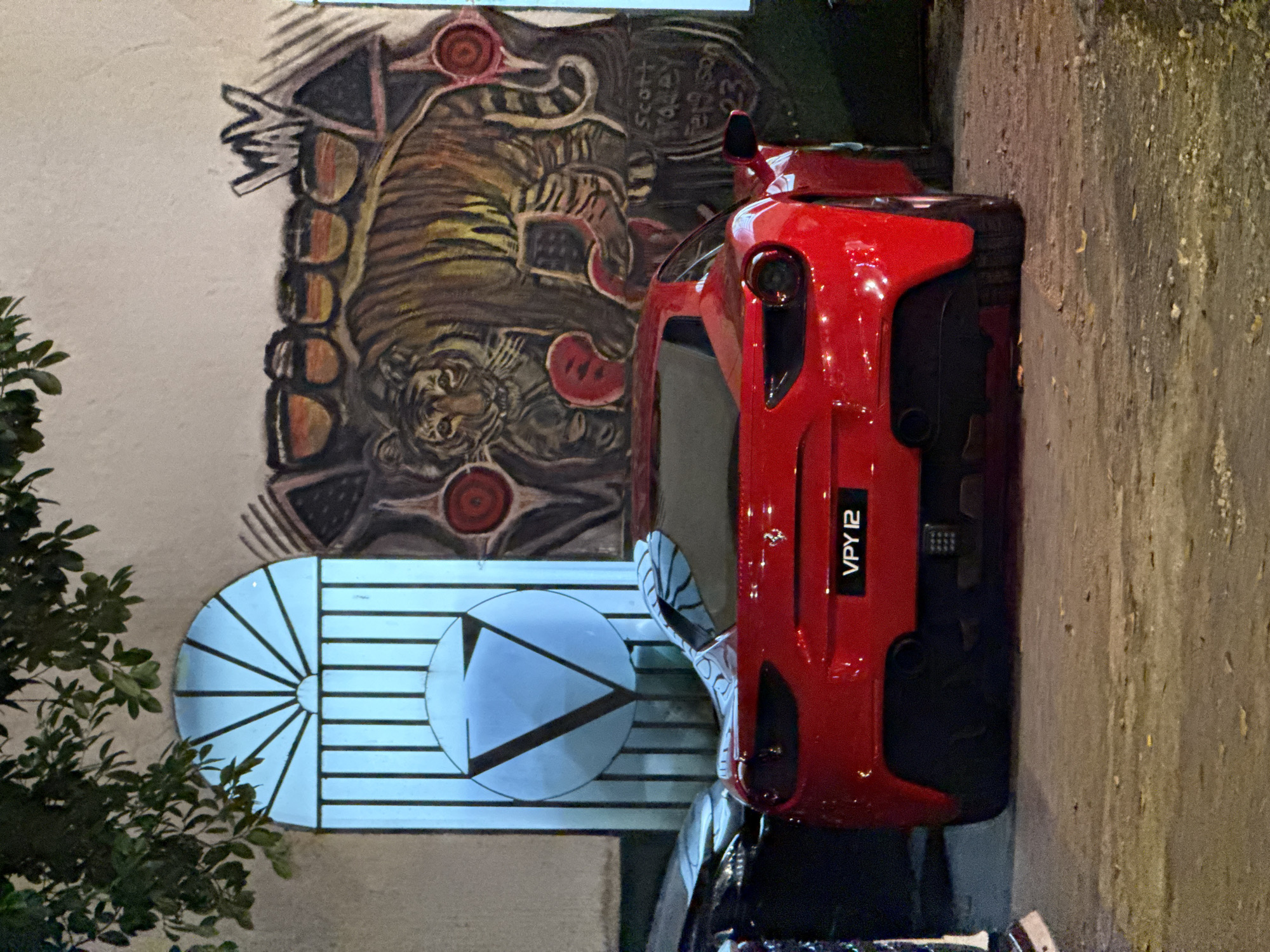 The image depicts a red Ferrari sports car parked on a street in Kuala Lumpur, Malaysia. The car has a license plate that reads "VPY 12." Behind the car is a wall with a mural depicting a tiger, along with other geometric shapes and what appears to be some writing. To the left of the mural, foliage partially obscures the view. The street pavement is visible to the right of the car. The light suggests it is nighttime.