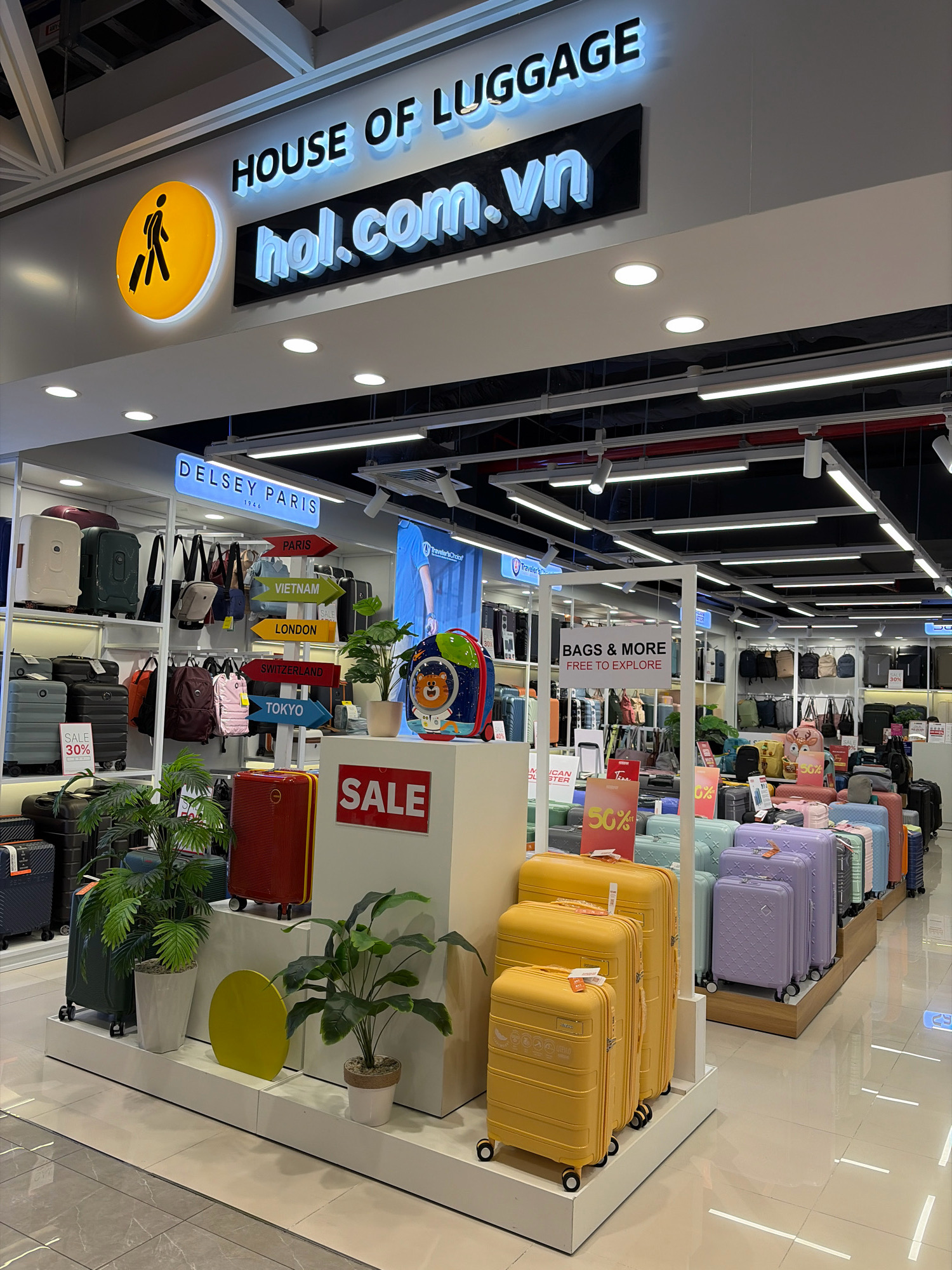 This image depicts the interior of a "House of Luggage" store, identified by signage, likely located in Da Nang, Vietnam. The store prominently displays a wide variety of suitcases and travel bags. In the foreground, several yellow suitcases of varying sizes are arranged on a white platform, with a green potted plant to their left and another to their right. Behind these, a prominent red suitcase stands out, positioned next to a white display box with "SALE" written in red. Further into the store, rows of colorful suitcases, including purple and light blue, are visible on tiered wooden shelving. Various brands and styles of luggage, including backpacks and duffel bags, are displayed on racks and shelves throughout the store. A sign with directional arrows pointing to "PARIS," "VIETNAM," "LONDON," "SWITZERLAND," and "TOKYO" is also visible. The overall environment appears to be a retail space with bright overhead lighting.