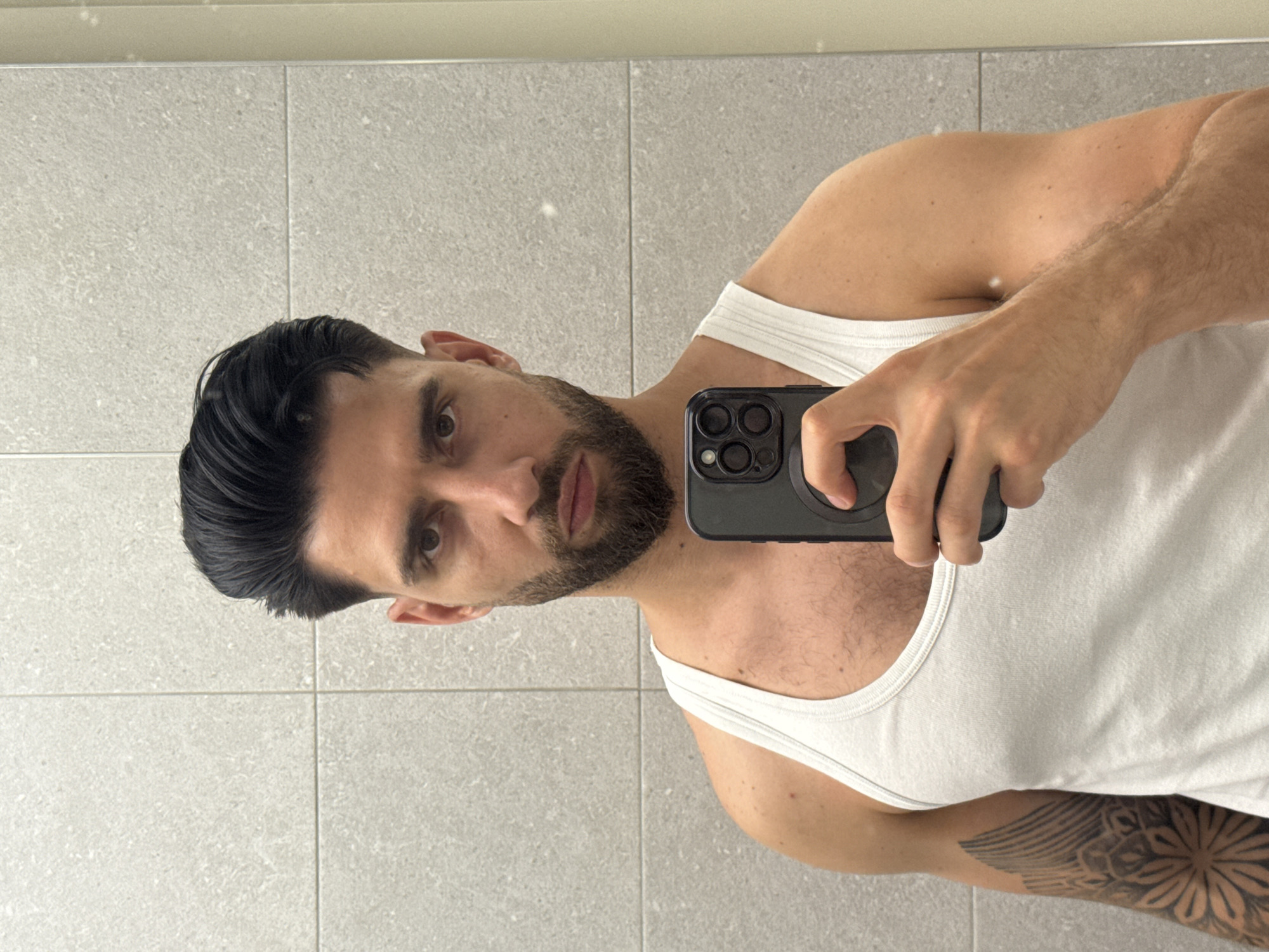 A man is taking a selfie in a bathroom. He has dark hair, a beard, and is wearing a white tank top. A tattoo is visible on his arm. He is holding a black iPhone. The background shows a wall of gray tiles. The location, Sofia, Bulgaria, cannot be determined from the image.