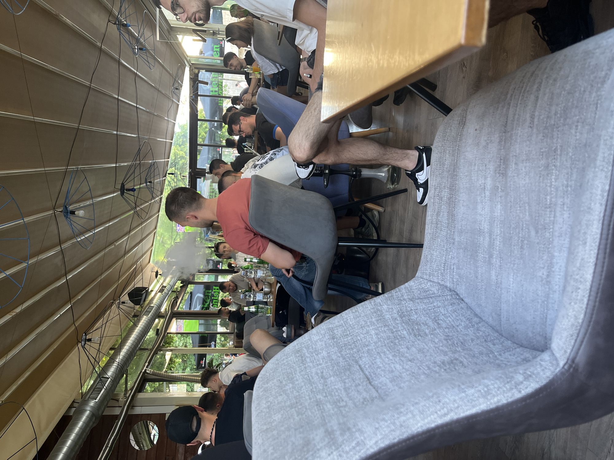 The image shows a crowded indoor space in Unknown, Unknown, possibly a cafe or lounge, filled with people seated at tables. The space has a partially glass ceiling. Several individuals are smoking, with visible plumes of smoke in the air. People are dressed casually. Tables are covered with drinks and other objects. The room is filled with chairs of different styles.