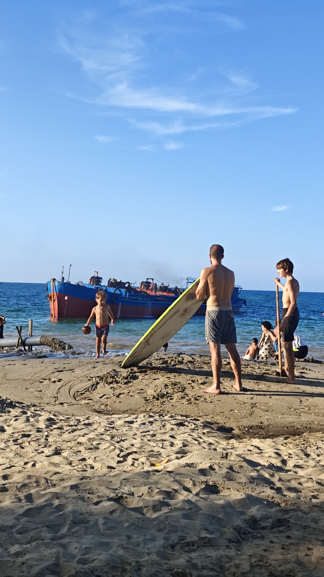 A boy holding a coconut is running out of the ocean, onto a beach in the city of Đà Nẵng, Vietnam. Behind him in the water, is a large blue and red industrial ship. On the sand, a man with his back to the viewer is holding a long, yellow surfboard. Another boy stands nearby, holding a long stick. In the background, more people are on the beach. The sky is blue and partially clouded. The sand is dark and light-colored.