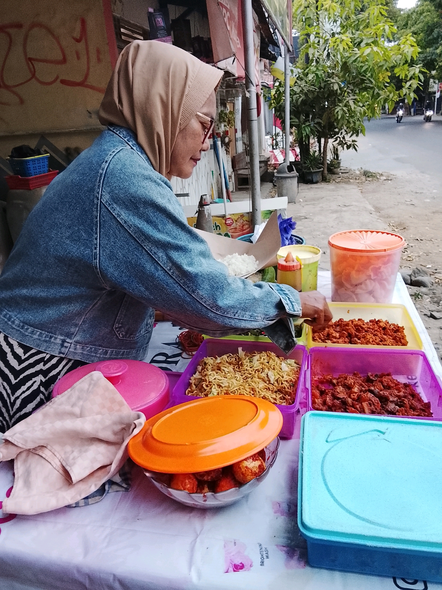 A woman wearing a beige hijab and a denim jacket stands behind a food stall in Slawi, Indonesia. She is serving food from various containers laid out on a table covered with a white tablecloth. In front of her are several food trays filled with what appears to be fried noodles, chopped meat, and other dishes. To her right, a large orange container holds snacks or crackers, and a yellow container holds sauce or condiments. The background shows a street scene with trees and passing motorcycles, indicating an outdoor urban environment. The woman's action suggests she is in the process of serving a customer or preparing food for sale.