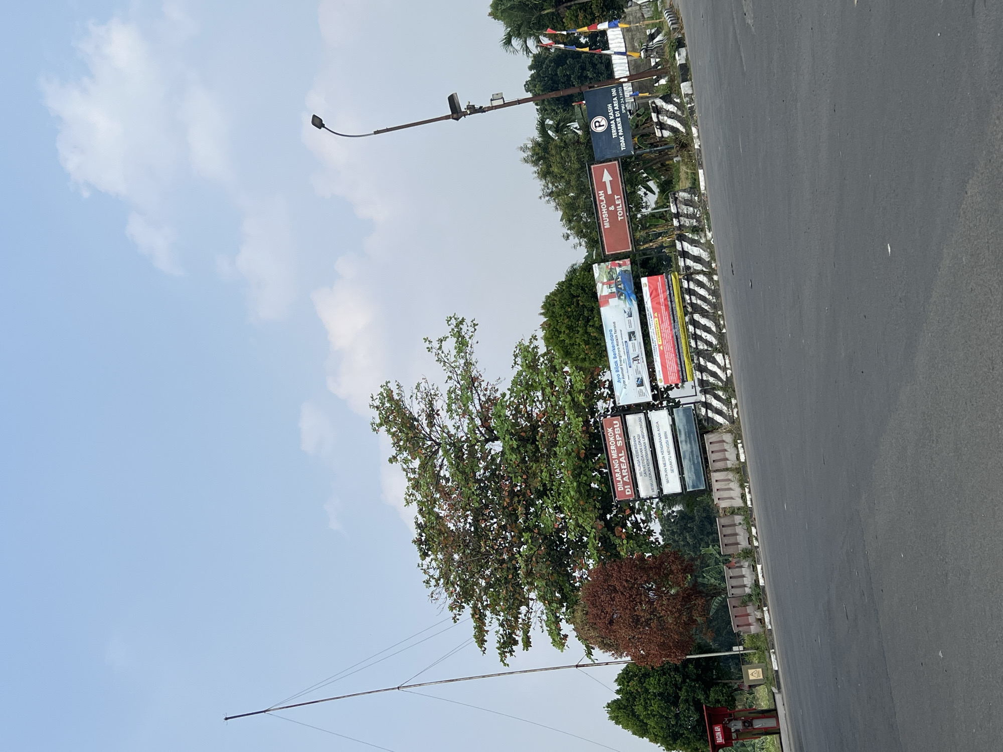 In the city of Gunung Putri, Indonesia, a scene unfolds on a wide, grey asphalt road with a clear blue sky above. A prominent, tall, thin pole with wires attached stretches upwards on the left side of the image. In the middle ground, a large tree with green and reddish-brown leaves stands out against a backdrop of smaller green trees. Several billboards are visible behind the tree, displaying text and images. One billboard prominently states "DILARANG MEROKOK DI AREA SPBU" (No Smoking in the Gas Station Area). Another sign reads "MUSHOLAH & TOILET" with an arrow pointing to the right. A parking sign with a "P" symbol is also present, indicating "TIDAK PARKIR DI AREA INI" (No Parking in this Area). In the foreground, concrete barriers with black and white stripes line the side of the road. To the far left, a small red structure with the text "RACUN API" (Fire Extinguisher) is visible. The environment appears to be a roadside near a gas station.