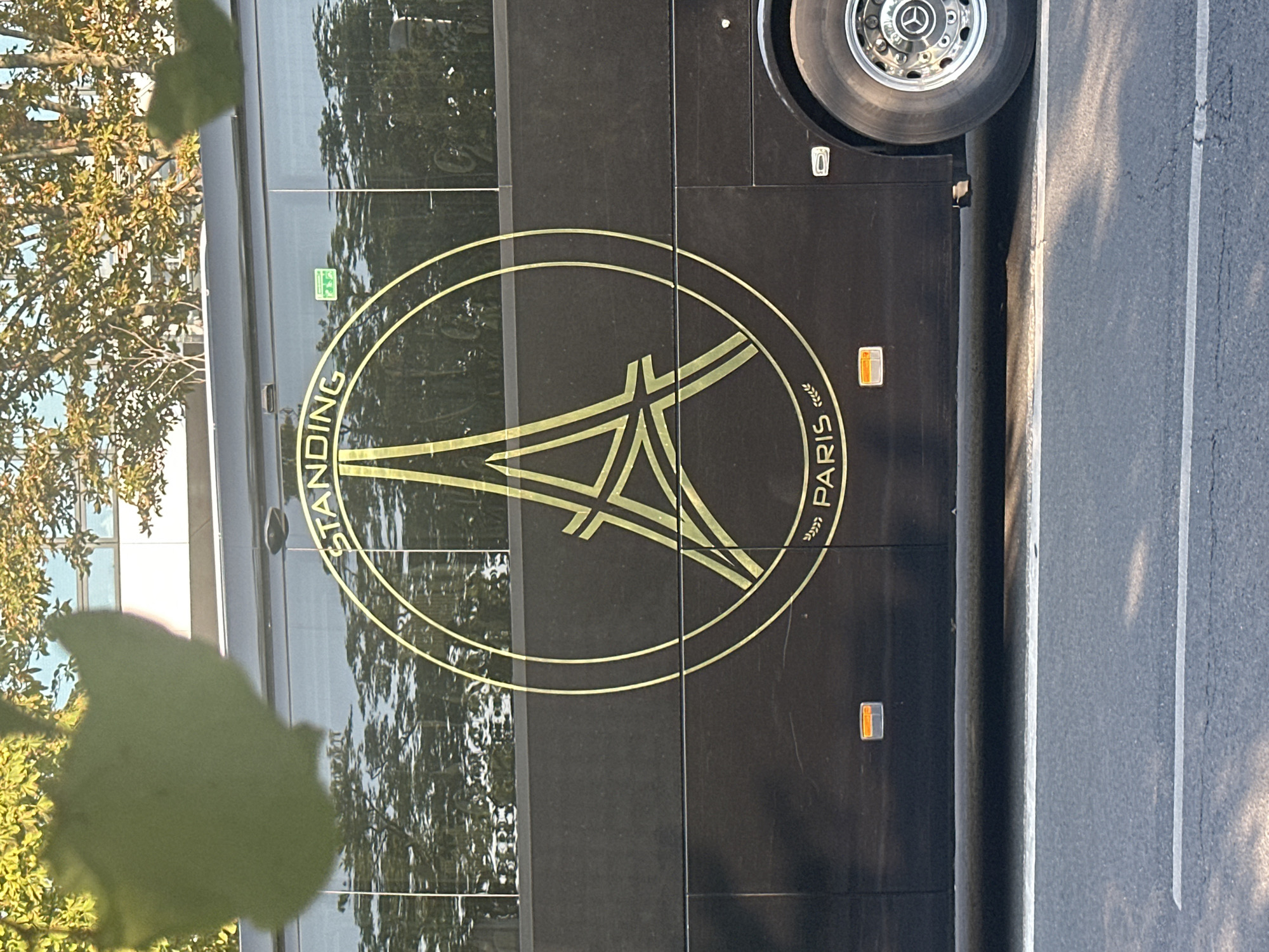 The image shows a side view of a black bus with a gold circular logo featuring an Eiffel Tower illustration. The text "STANDING PARIS" is visible within the logo. The bus is parked on a street, with reflections of trees and buildings visible in its windows. The lower part of the image shows the road surface and a white lane marking. The scene appears to be in an urban environment, likely in Paris, France, given the logo. The bus has a visible tire with a silver rim and a Mercedes-Benz logo. There are no people visible in the image. The lighting suggests it is daytime.
