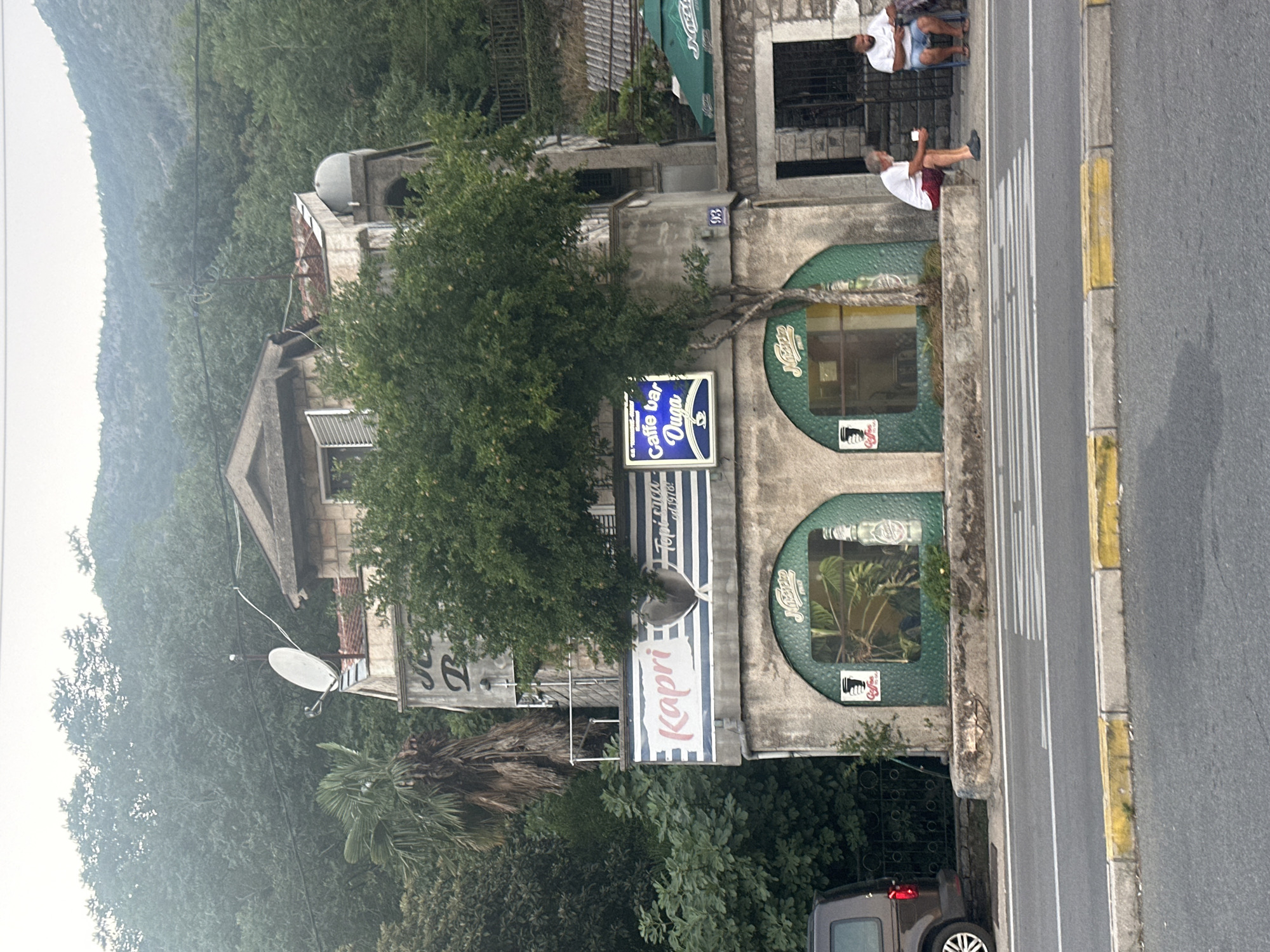 In Jošice, Montenegro, a two-story building with a rough, weathered facade stands at the foot of a steep, tree-covered hill. The building appears to house a cafe or bar, indicated by signage that includes "Kapri" and "Caffe bar Duga." The lower level features two arched window openings, one displaying an image of tropical plants and a bottle, the other a coffee cup icon. A mature tree grows in front of the cafe, its branches reaching towards the upper story. Above the cafe, a balcony with a railing is visible, and further up the building, a satellite dish is mounted. Two men are seated on a curb in front of the building; one is an elderly man with white hair and a beard, wearing a white t-shirt and red shorts, holding a white cup. The other man, with darker hair and a beard, wears a white t-shirt and blue shorts, and is also holding a phone or small object. A crosswalk is present on the asphalt road in front of the building, with yellow and white lines marking its edges. A dark-colored van is parked to the left of the frame. The environment is lush and green, with dense vegetation covering the hillside behind the building. The sky is overcast, suggesting a cloudy day.