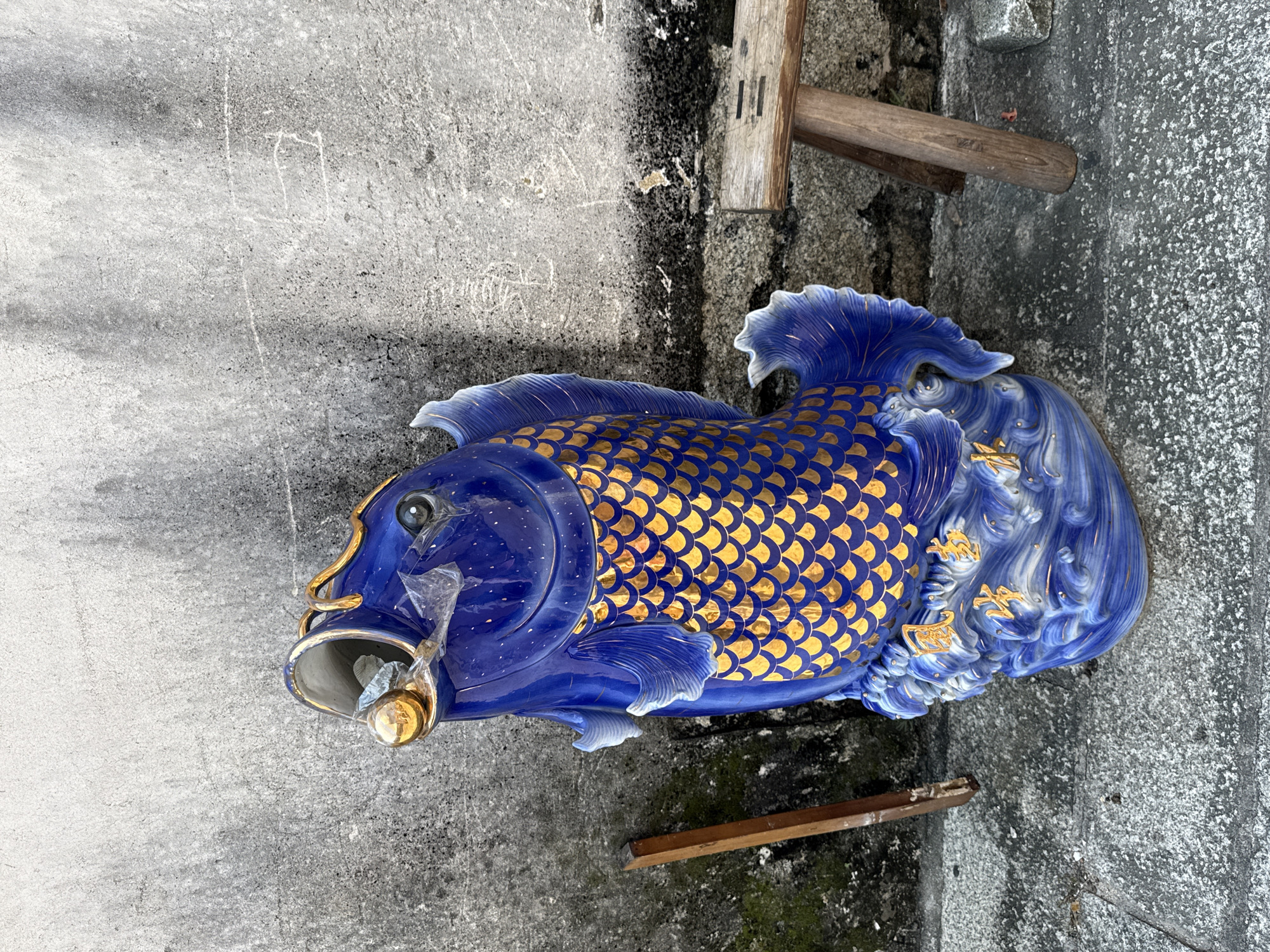 A vibrantly blue ceramic fish sculpture with gold scale detailing stands on a textured gray concrete surface in front of a rough, gray concrete wall. The fish has its mouth open, with a gold orb held inside, and appears to be perched on a base decorated with swirling blue and white waves, also accented with gold. A wooden object, possibly a stool or part of a structure, is positioned to the right of the sculpture, and another wooden piece is leaning against the wall to the left. The overall environment suggests an outdoor urban setting, likely in Longgang District, China, given the architectural style of the wall and the concrete paving. There are no people visible in the image.