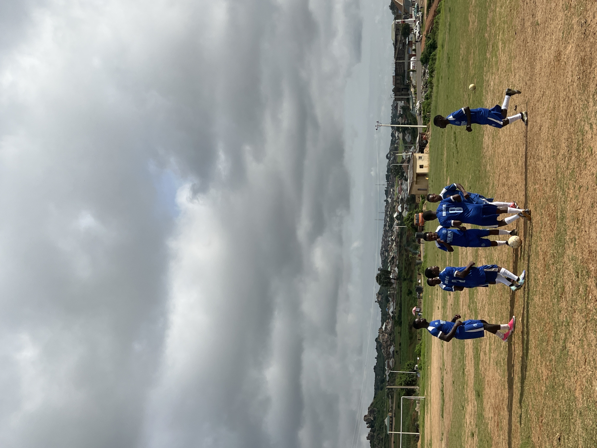 The image depicts a group of young men in blue soccer uniforms with white accents on a dry, grassy soccer field in Jos, Nigeria. The sky is overcast with heavy, gray clouds. In the background, a sprawling settlement with various buildings, some with corrugated metal roofs, is visible on a hillside dotted with rocky outcrops and scattered trees. A soccer goalpost is present on the left side of the frame. The players are engaged in a practice session, with some standing in a huddle and others in motion. One player is kicking a soccer ball. The overall impression is that of a casual soccer training session taking place in a semi-urban environment under a moody sky.