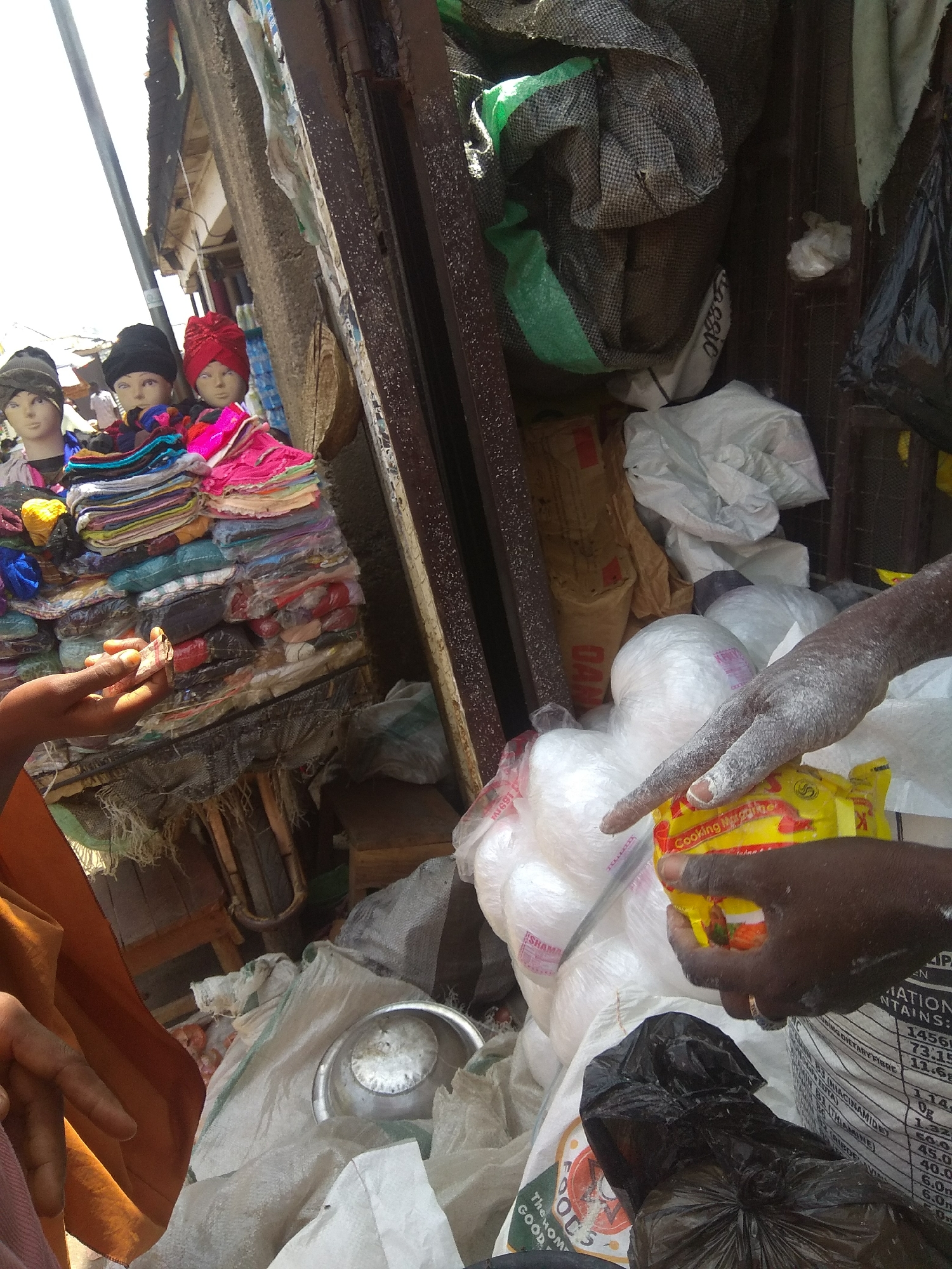 This media file captures a bustling market scene in Jalingo, Nigeria. The foreground is dominated by the hands of a person holding a small packet labeled "Cooking Margarine," with their other hand reaching towards a pile of stacked goods. A variety of items are visible, including textiles, what appear to be bags of flour or grains, and plastic-wrapped goods. Mannequins wearing headscarves are also present, displayed alongside neatly folded fabrics. The setting is an open-air market, suggested by the sunlight and the arrangement of goods on what seems to be makeshift stalls. The overall impression is one of commerce and daily life in a vibrant Nigerian marketplace. The lighting suggests it is daytime, likely under a bright sun. The text visible on the packet and on some of the bags provides clues about the products being sold, with specific nutritional information and brand names like "DAN" and "SHAM." The presence of money being exchanged also indicates a transaction is underway.