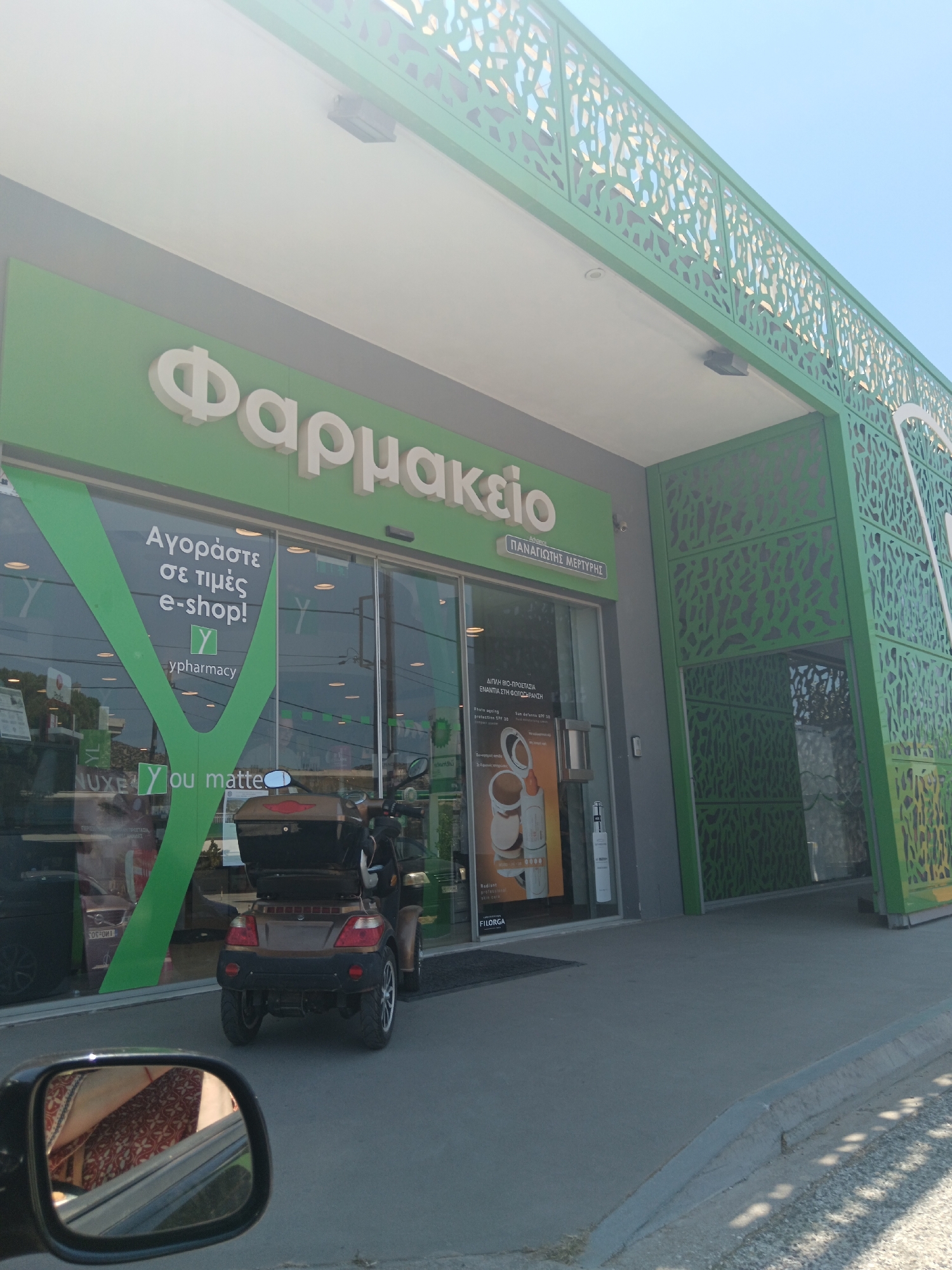 The image depicts the exterior of a pharmacy with green architectural elements. The pharmacy's name, "Φαρμακείο," is prominently displayed in white, three-dimensional lettering on a green background above the entrance. A smaller sign below reads "ΠΑΝΑΓΙΩΤΗΣ ΜΕΡΤΥΡΗΣ". The entrance features large glass doors, and a green stylized "Y" graphic is visible on the left door, along with text that appears to be "Αγοράστε σε τιμές e-shop!".
Outside the pharmacy, a three-wheeled mobility scooter with a dark brown body and a black storage compartment is parked on the sidewalk. To the left, a car's side mirror is visible in the foreground, reflecting a patterned fabric interior. The ground surface is paved concrete. The sky is clear and blue. The overall environment suggests a sunny day in a commercial area. The location is identified as Agia Marina, Greece.
