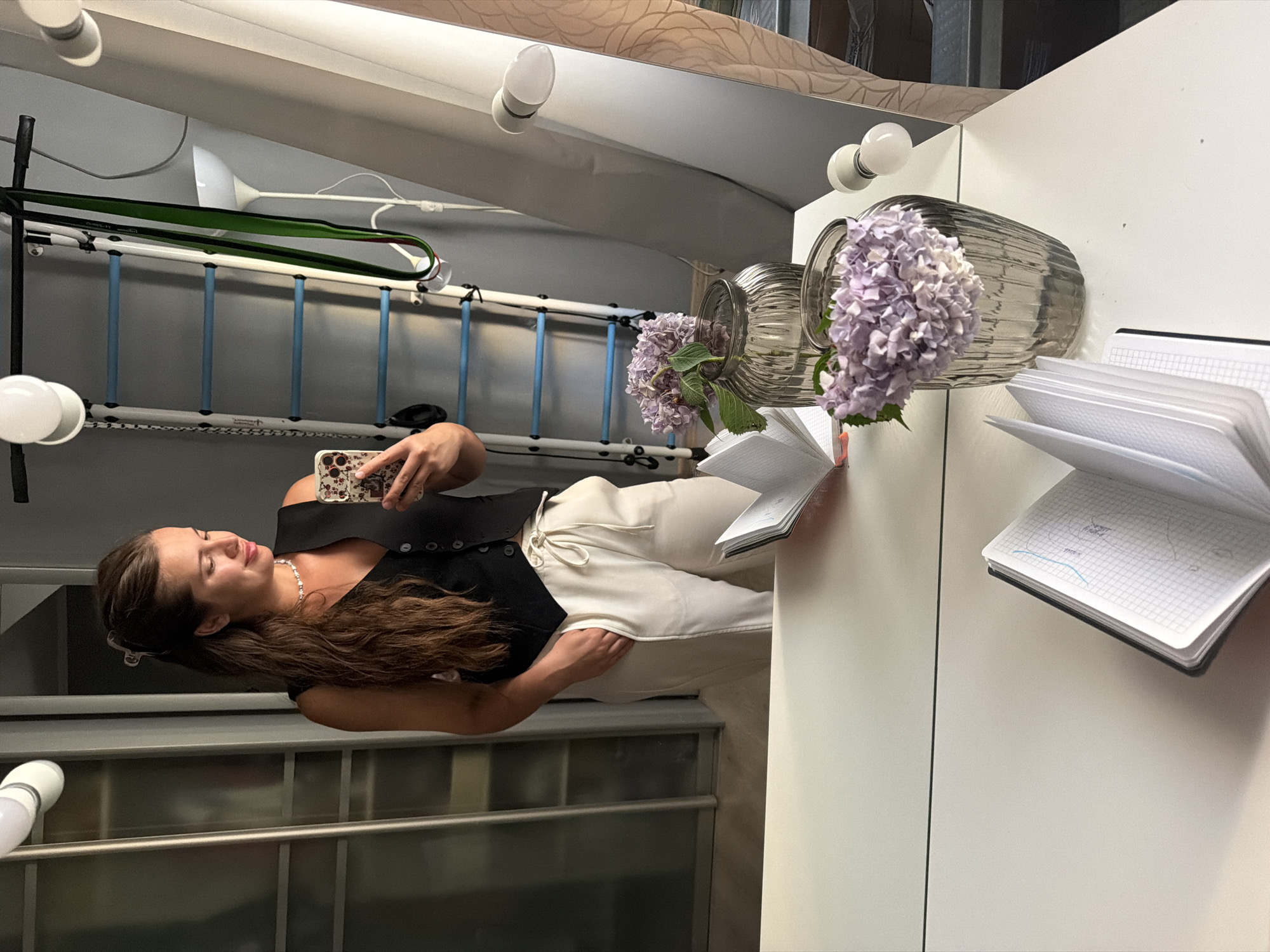 A woman is taking a selfie with her phone in a brightly lit room. She is wearing a black top and white pants and has long brown hair. She is standing in front of a white counter with a vase of purple hydrangeas and an open notebook. In the background, there is a ladder and some pipes. The room appears to be a modern, indoor space, possibly a studio or a home. The woman has a relaxed expression on her face. The time of day is not discernible from the image.