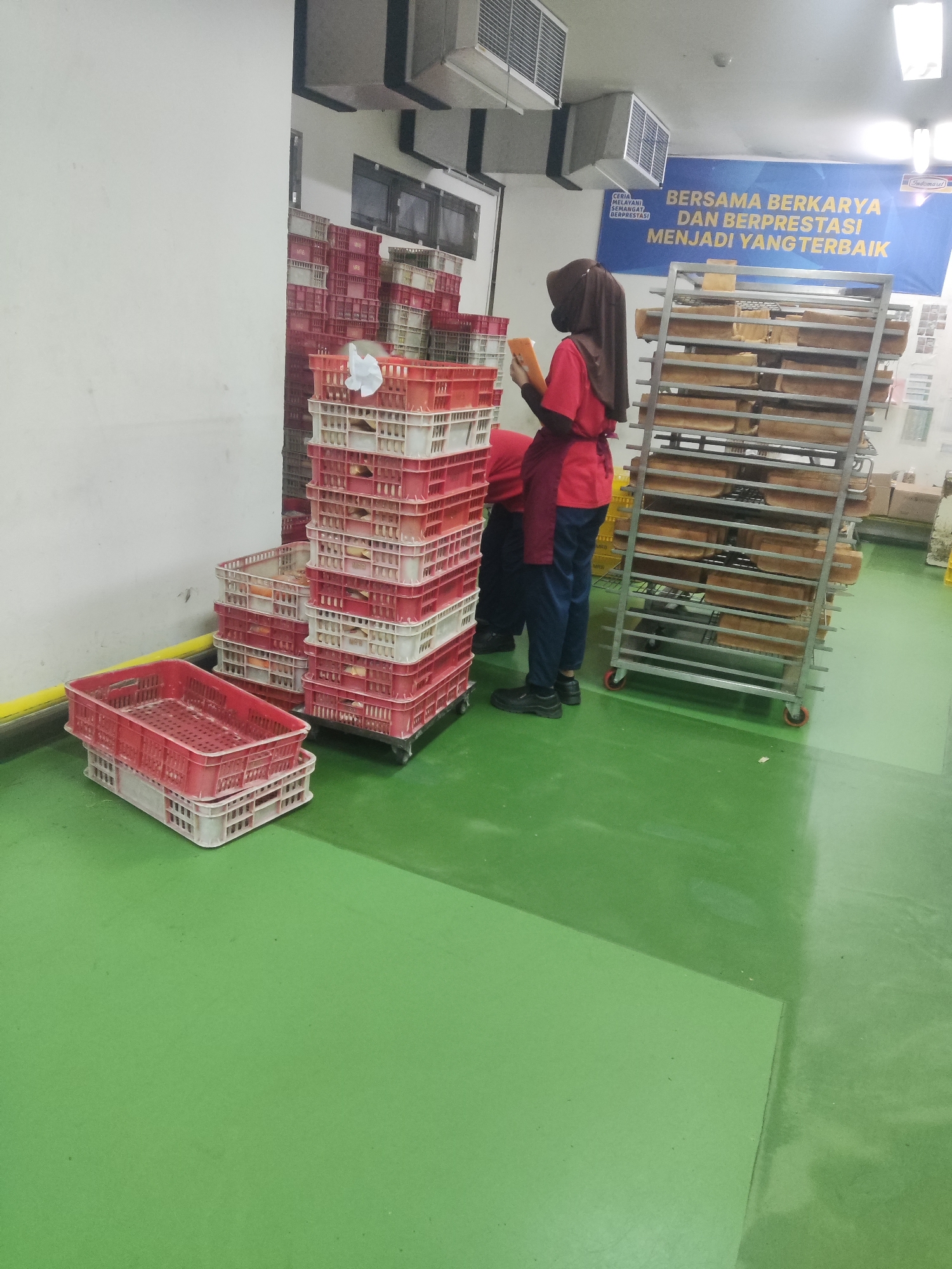 The image shows a food production or packaging facility. In the foreground, there are stacks of red and white plastic crates. Two individuals are visible; one, wearing a brown hijab and a red shirt, is holding an orange object. The second individual, wearing a red shirt and dark pants, is standing behind the first. In the background, there is a large metal rack filled with baked goods, likely bread. A blue banner with text is displayed on the wall. The floor is green. The scene appears to be in a commercial or industrial setting within Trihanggo, Indonesia.