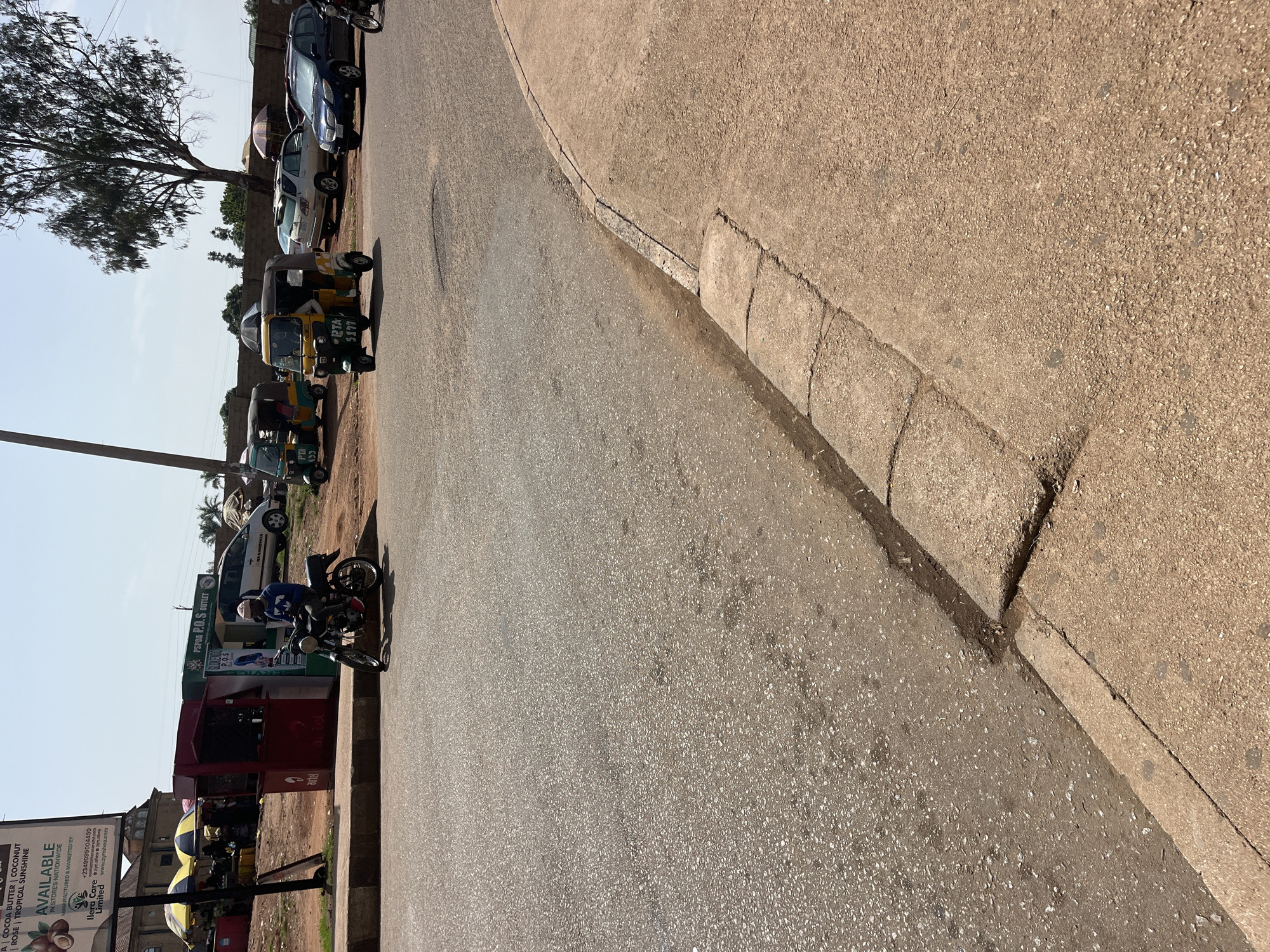 The image captures a street scene in Gura Top, Nigeria, likely near a commercial area given the presence of stalls and a busy road. In the foreground, a motorcycle with a rider wearing a blue shirt, dark pants, and a cap is parked on the asphalt road. The rider is looking towards the left. Further down the road, a yellow and green motorized tricycle, commonly known as an "okada" or "kekeke," is visible. It has "PTA 433" and "PTA 5177" written on it. Other vehicles, including a white SUV and a blue sedan, are parked or driving on the road in the background. Several market stalls with awnings are set up along the side of the road, suggesting a marketplace. The environment appears dry and dusty, with a few trees scattered in the background. The sky is clear and bright, indicating a sunny day.