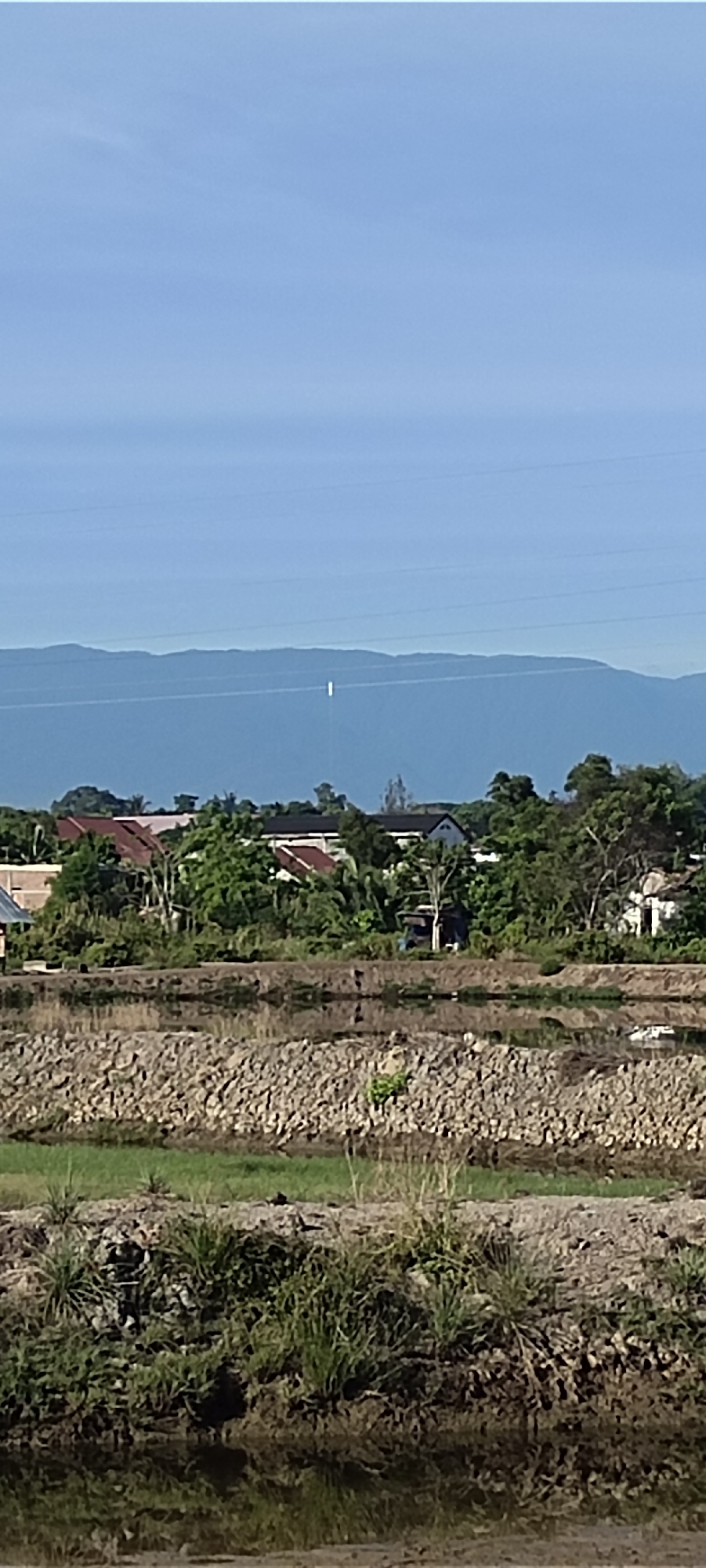 The photo depicts a partially submerged landscape in Lhokseumawe, Indonesia, under a clear, pale blue sky with distant mountains. Overhead power lines run horizontally across the upper portion of the frame. The foreground features flooded agricultural land with patches of vegetation and rocky banks. Behind this, a line of trees and houses with red and white roofs forms a mid-ground barrier. The mountains are in the distance. A single bright white object is visible between the mountains and powerlines.