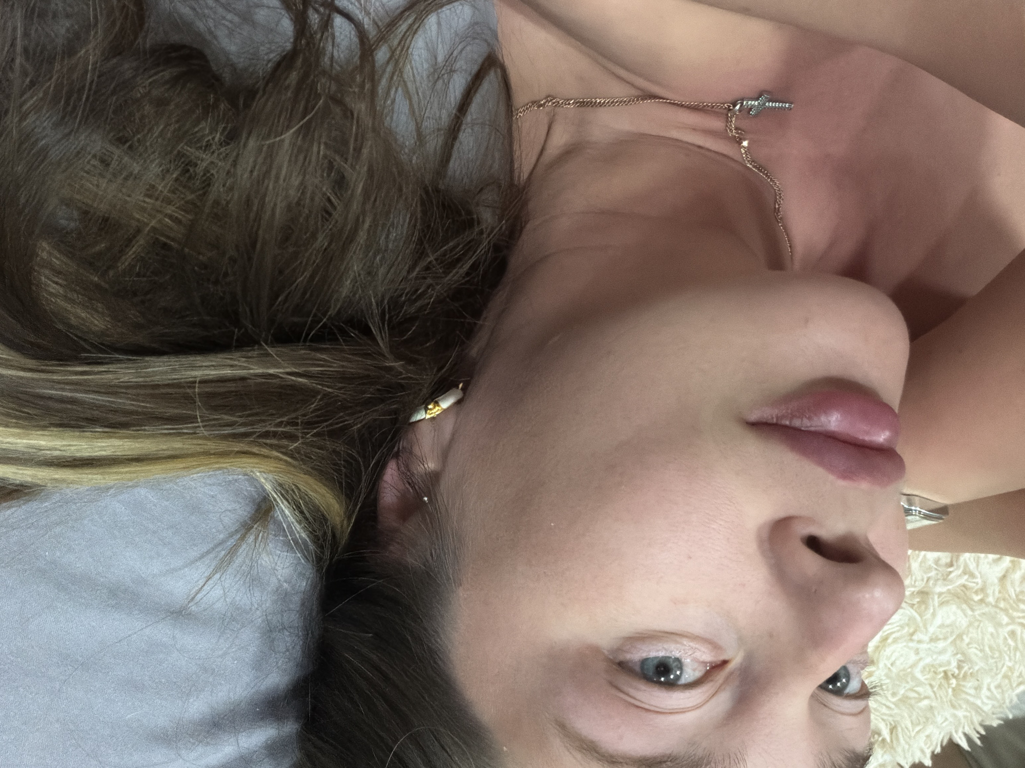 A close-up, angled portrait of a woman lying on her back. She has fair skin, blue eyes, and light brown hair with blonde highlights. She is wearing a gold necklace with a cross pendant and a gold earring. She appears to be lying on a grey pillow and a white, fluffy surface. The location is Chaikovsky, Russia is not discernible from the image.