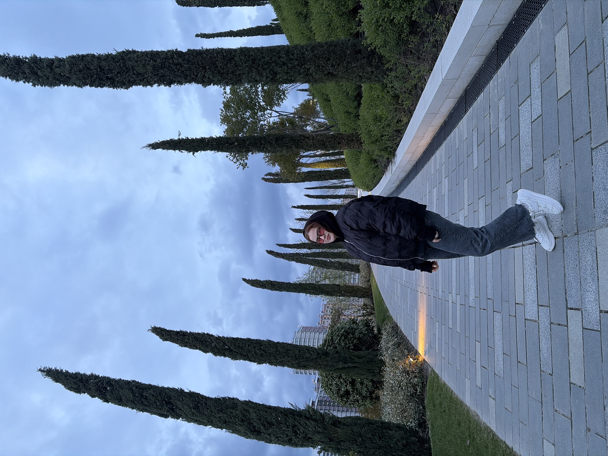 An outdoor, eye-level shot captures a person standing on a paved pathway under an overcast sky in what appears to be a modern urban park or promenade in Krasnodar, Russia.
The central subject is a person, likely young, standing casually with their right hand in a pocket, facing the camera. They are dressed in a black hooded jacket, dark grey jeans, and white athletic sneakers, and wear red-tinted sunglasses.
The pathway is constructed from rectangular grey paving stones. To the person's right, a low concrete barrier with a dark grate runs alongside the path. Beyond this barrier, rows of tall, slender evergreen trees, resembling cypresses, line the walkway, creating a strong sense of perspective. Further back, additional greenery and modern multi-story buildings are visible in the background. To the left of the path, a manicured green lawn and more dense foliage are present. A warm, possibly artificial, orange light illuminates a section of the paved path.
The sky is a uniform light grey, filled with clouds, indicating an overcast day. The overall lighting suggests it might be late afternoon or early evening. There is no visible text in the image.