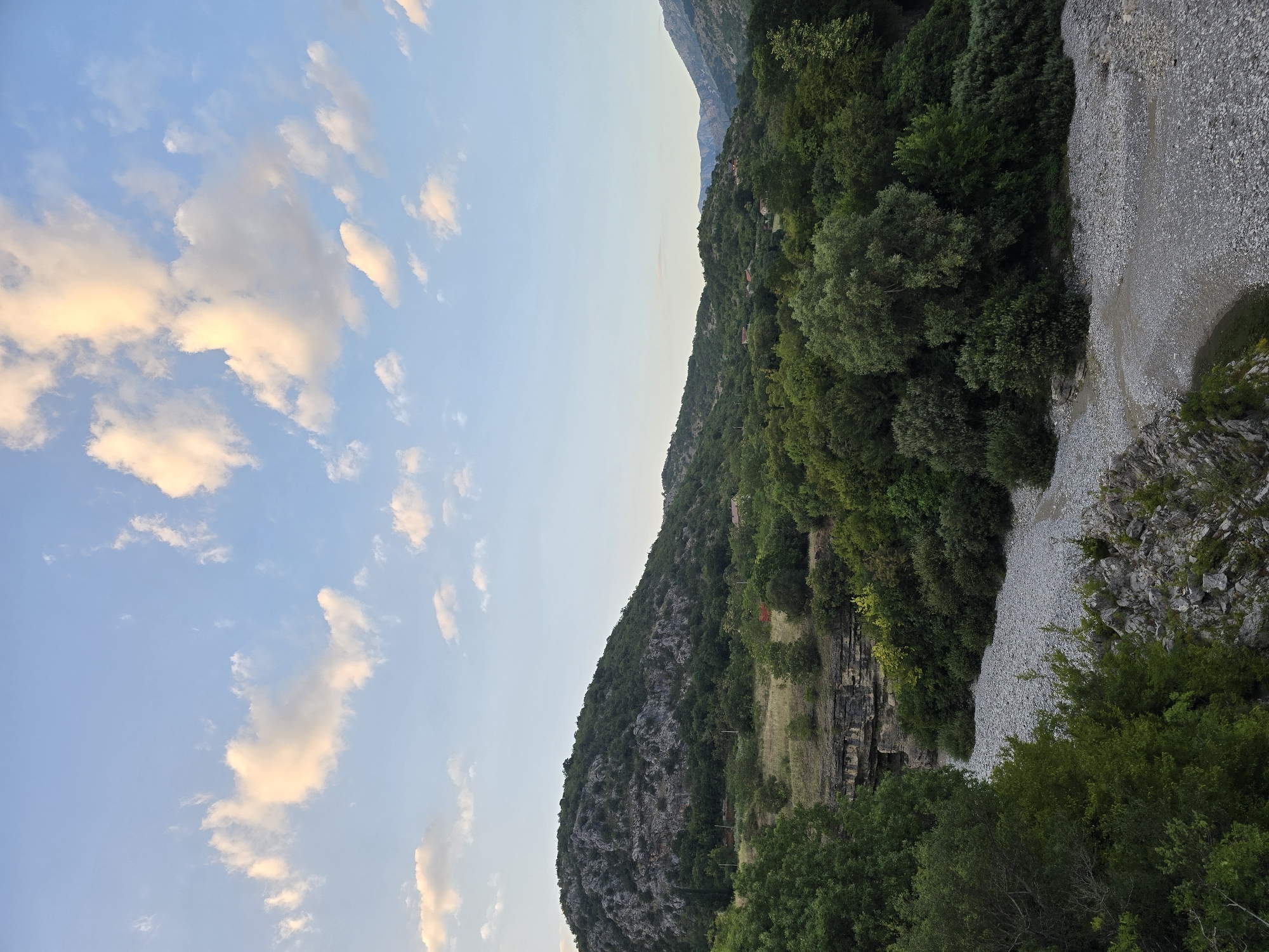 The image depicts a natural landscape under a sky with scattered clouds. In the foreground, a dry riverbed filled with grey pebbles dominates the lower portion of the frame. Lush green trees and vegetation line the sides of the riverbed and extend up the slopes of rolling hills. A prominent rocky hill covered in dense green foliage occupies the middle ground, with hints of exposed rock faces. Further in the distance, more hills and mountains are visible, receding into a hazy blue horizon. The sky is a pale blue, with fluffy white and golden clouds, suggesting it might be late afternoon or early evening. The overall scene is serene and undeveloped, characteristic of a rural or wilderness setting. No people or man-made structures are clearly discernible in the image, apart from what might be very small, distant buildings amongst the trees on the hillsides.