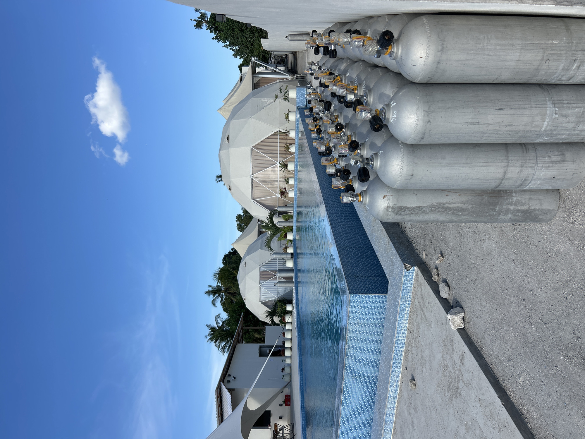 A row of silver scuba diving tanks are lined up along a white wall on a dirt and gravel ground. In front of the tanks is a blue-tiled swimming pool with clear blue water. Behind the pool are several white dome-shaped structures and a white building with palm trees in the background. The sky is a clear blue with a few scattered white clouds. The scene appears to be a resort or dive center on the island of Nusa Penida, Indonesia.