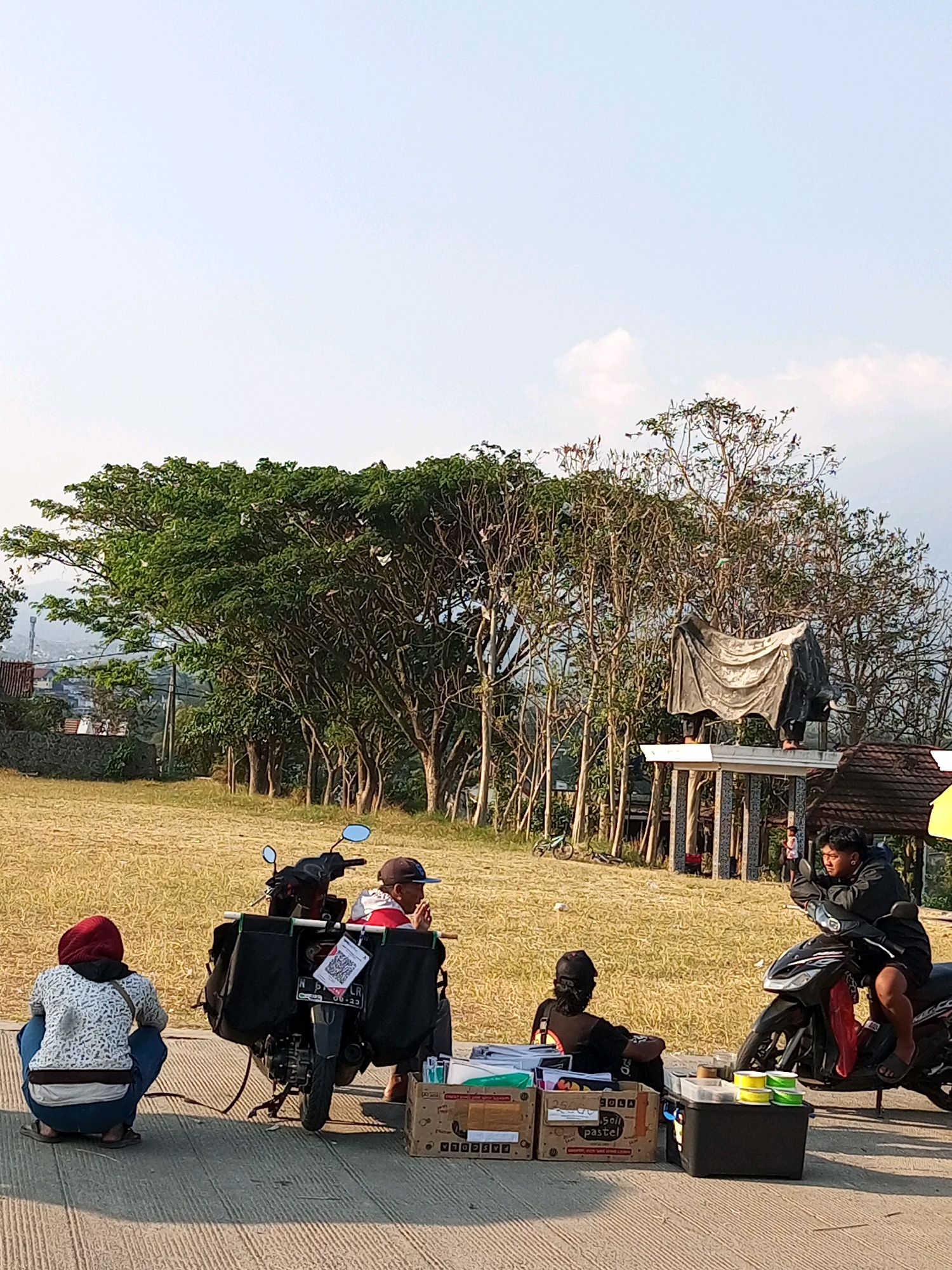 In Batu, Indonesia, a scene unfolds on a paved surface adjacent to a dry, grassy field. The sky is a pale blue with scattered clouds. In the foreground, three people are visible. To the left, a person wearing a red head covering and a patterned jacket squats facing away from the camera. Beside them, a motorcycle is parked, equipped with two large black saddlebags. A person wearing a red shirt and a baseball cap sits on the motorcycle, looking towards the right. In the center, a pile of boxes and containers sits on the ground. To the right, a person wearing a black jacket and shorts sits on another motorcycle, appearing to interact with someone. Behind them, a structure covered with a dark tarp stands, with several trees surrounding it. Further in the background, a hillside with buildings and vegetation is visible, along with a cell tower.