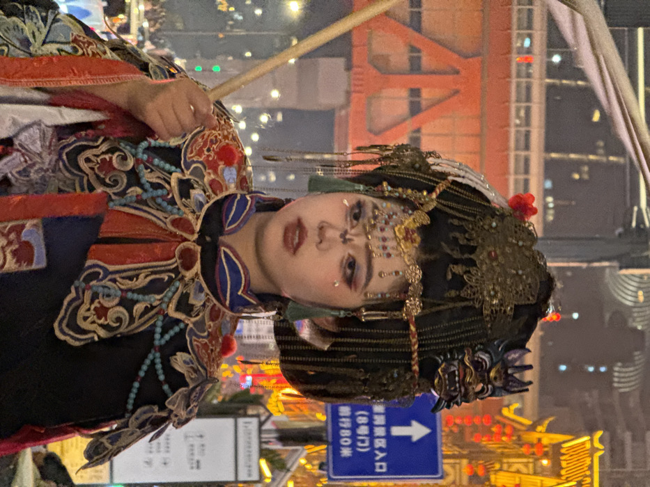 An individual, likely female, is depicted in a medium shot at night in the Yuzhong District of Chongqing, China, specifically near the Hongyadong Scenic Area entrance. The person faces slightly right, presenting a neutral expression, and is adorned in elaborate traditional Chinese attire and heavy stage makeup. The costume features a dark base garment with a high collar and a prominent, multi-layered embroidered shoulder piece (yunjian) in patterns of red, blue, gold, and white, accented with turquoise beads and small red pom-poms. The individual's dark hair is styled into a complex updo, embellished with intricate gold-colored metallic ornaments, red accents, green tassels, and a dark, fanged mask-like adornment on the right side of the head. Facial makeup includes defined brows, elaborate red and dark eyeshadow, stylized white markings extending from the inner eyes towards the forehead, a gold-colored decorative element on the forehead, and reddish-brown lipstick. The left hand holds a light-colored wooden staff or pole, angled upwards.
The background is a vibrant urban night scene. To the left, a large orange-red metal structural element, possibly part of a bridge or building, is visible, partially obscured. To the right, a blue rectangular sign with white text indicates "洪崖洞景区入口" (Hongyadong Scenic Area Entrance), "(8号门)" (Gate 8), and "前行80米" (Ahead 80 meters), confirming the location. Behind and to the right of the sign, multiple brightly illuminated structures with traditional Chinese architectural features are visible, decorated with numerous red lanterns and golden elements.