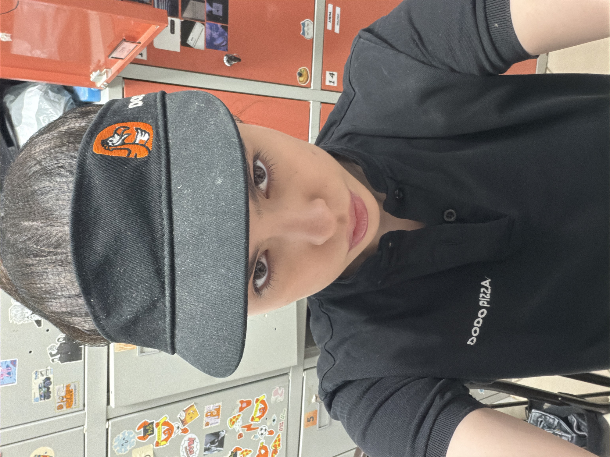 A young person wearing a black Dodo Pizza uniform, consisting of a polo shirt and visor, is looking at the camera. The shirt has "DODO PIZZA" embroidered on the left breast. The visor has an orange logo on the front. The person is in a room with gray lockers with stickers on them and orange lockers behind. This is likely a Dodo Pizza location in Chaikovsky, Russia, given the branding on the uniform.
