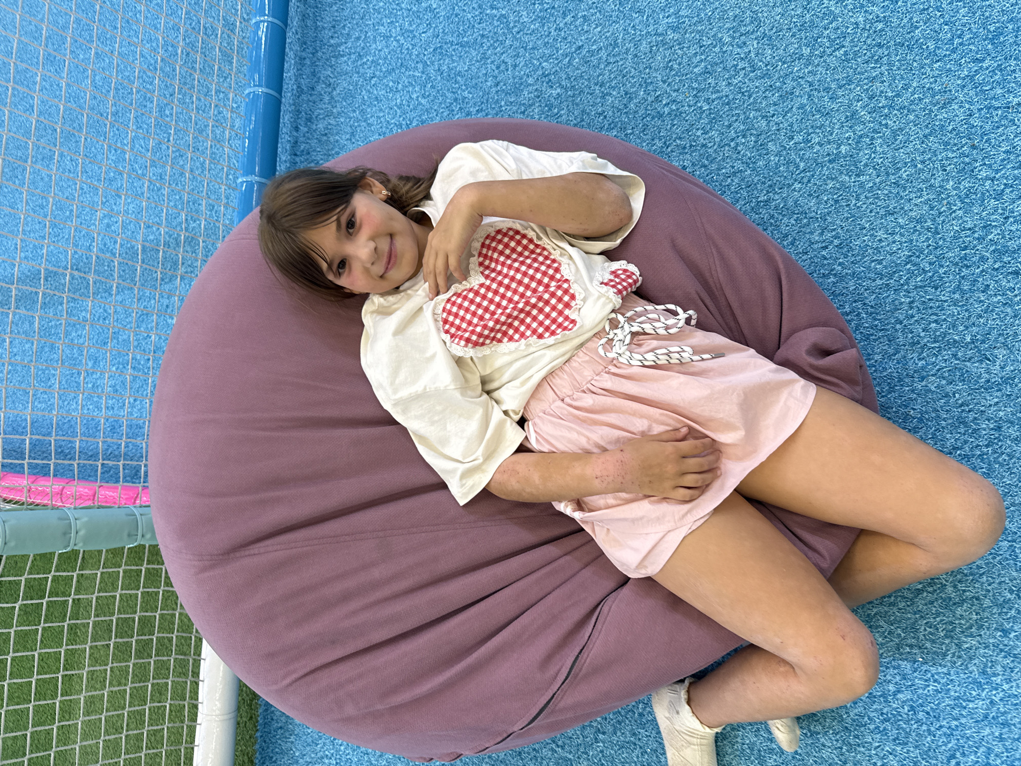 A young girl is lying on a large, plum-colored beanbag chair. She is wearing a light-colored t-shirt with a red and white checkered heart appliqué on the chest and a pale pink skirt. She has dark hair and is smiling at the camera. Her legs are crossed and her arms are resting on her lap. The beanbag is on a bright blue, textured floor. To the left of the beanbag, there is a section of green artificial turf visible, enclosed by a white net with blue and pink bars. This suggests the setting might be an indoor play area or gym. The lighting appears to be even and bright, indicating it's likely daytime, possibly indoors. The girl's expression is happy and relaxed. There is no visible text in the image.