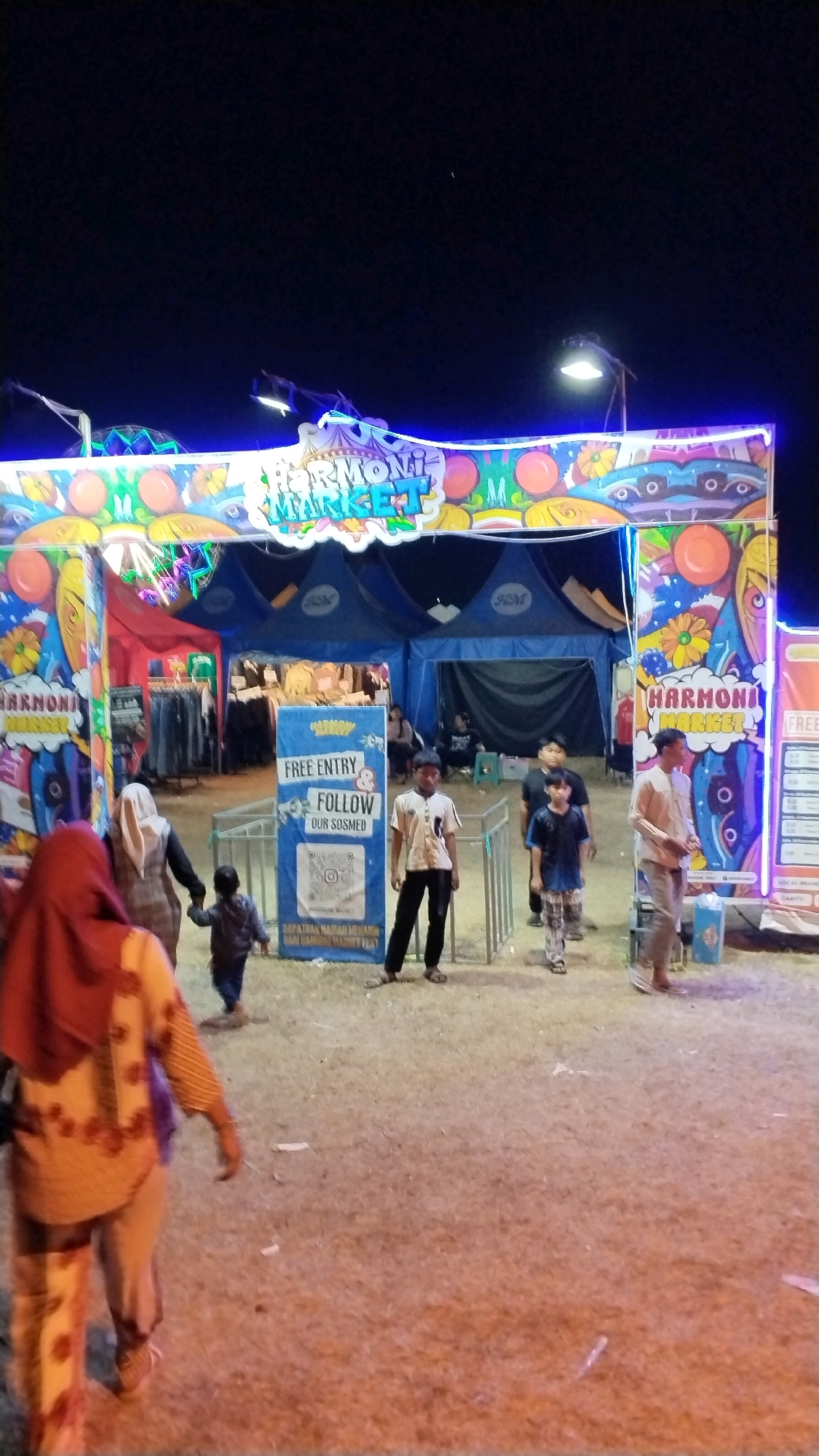 In Canggu, Indonesia, a lively night market scene unfolds under the glow of neon lights. A vibrant, arched entrance adorned with the words "HARMONI MARKET" in colorful, bubbly letters serves as the gateway to the event. The archway is decorated with stylized floral motifs and orange circular shapes.
Beneath the arch, several blue tents are visible, suggesting stalls or vendors. In front of the entrance, a signpost with "FREE ENTRY" and "FOLLOW OUR SOSMED" with a QR code indicates promotional information.
The ground is covered with dry, sandy earth, typical of an outdoor venue. Several people are present, including a woman in a red hijab and a patterned yellow and orange top in the foreground, walking away from the camera. To her left, a person in a brown hijab walks hand-in-hand with a young child.
Other individuals are seen near the entrance: a young boy in a light-colored shirt and dark pants stands to the right of the "FREE ENTRY" sign. Further back, a man in a light-colored shirt and pants is walking through the entrance. Other figures are visible in the background, appearing to be browsing the stalls or socializing. The overall atmosphere suggests a bustling community gathering or festival.