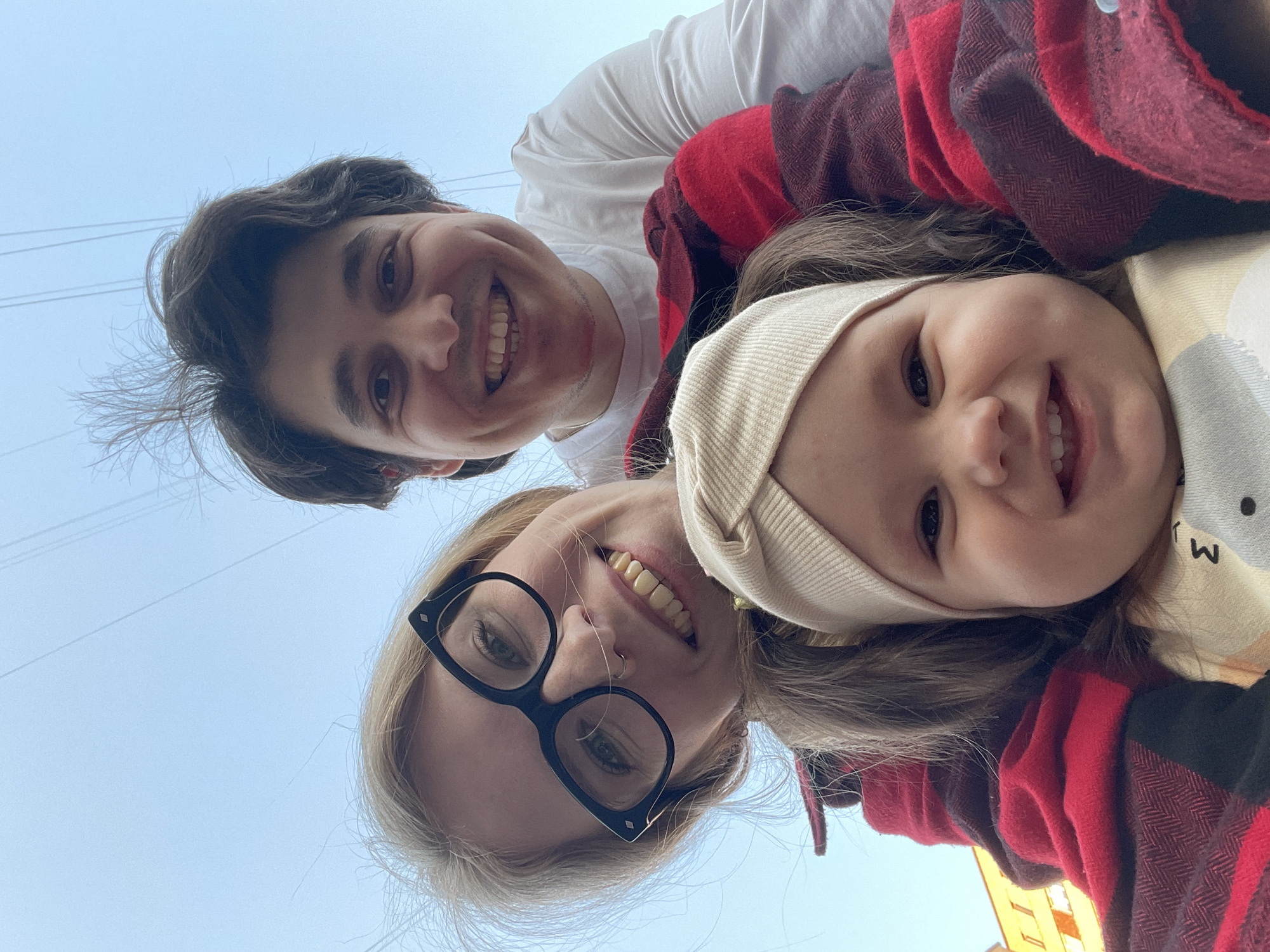 A photo taken looking upward shows three smiling people: an adult male in a white t-shirt, a woman with blonde hair and large black glasses, and a young child wearing a beige hat. The man is at the top of the frame, the woman at the bottom, and the child is between them, partially obscured by a red and black plaid jacket. The background is a pale blue sky with power lines visible near the top left. The bottom right corner shows a glimpse of a yellow building. The scene suggests an outdoor setting, possibly in Izhevsk, Russia, given the architectural style and background details. The photo appears to be a candid snapshot capturing a family moment.