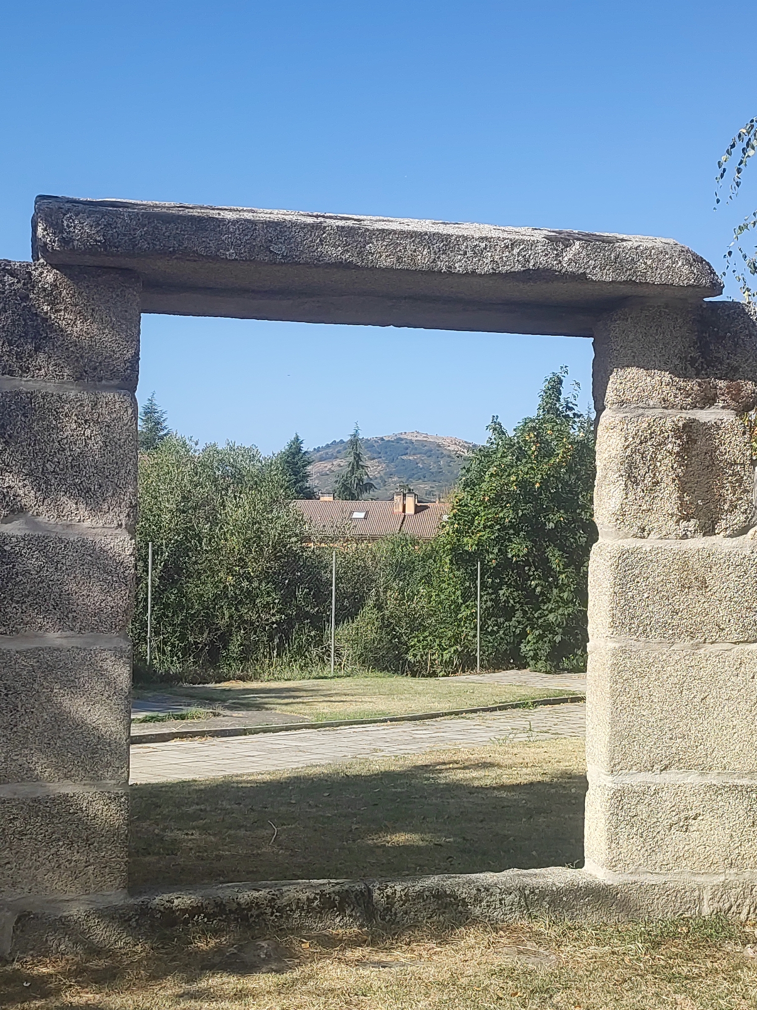 The image depicts a stone archway framing a view of a residential area and a distant hill. The archway is constructed from large, rough-hewn granite blocks, with a thick lintel spanning the top. Through the arch, a lush green canopy of trees and bushes is visible, partially obscuring a building with a red-tiled roof. Beyond the greenery, a hill rises in the background under a clear blue sky. The foreground shows dry grass and a paved pathway. The scene appears to be an outdoor setting in a town or village.