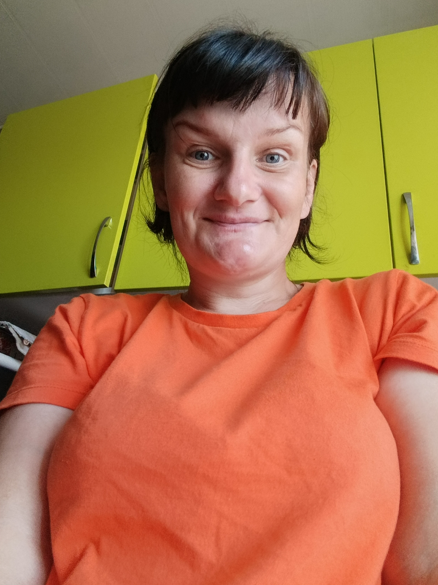 A woman is taking a selfie in what appears to be a kitchen. She has short brown hair with bangs and is wearing an orange t-shirt. Behind her are bright yellow kitchen cabinets with silver handles. The location is Unknown, Unknown.