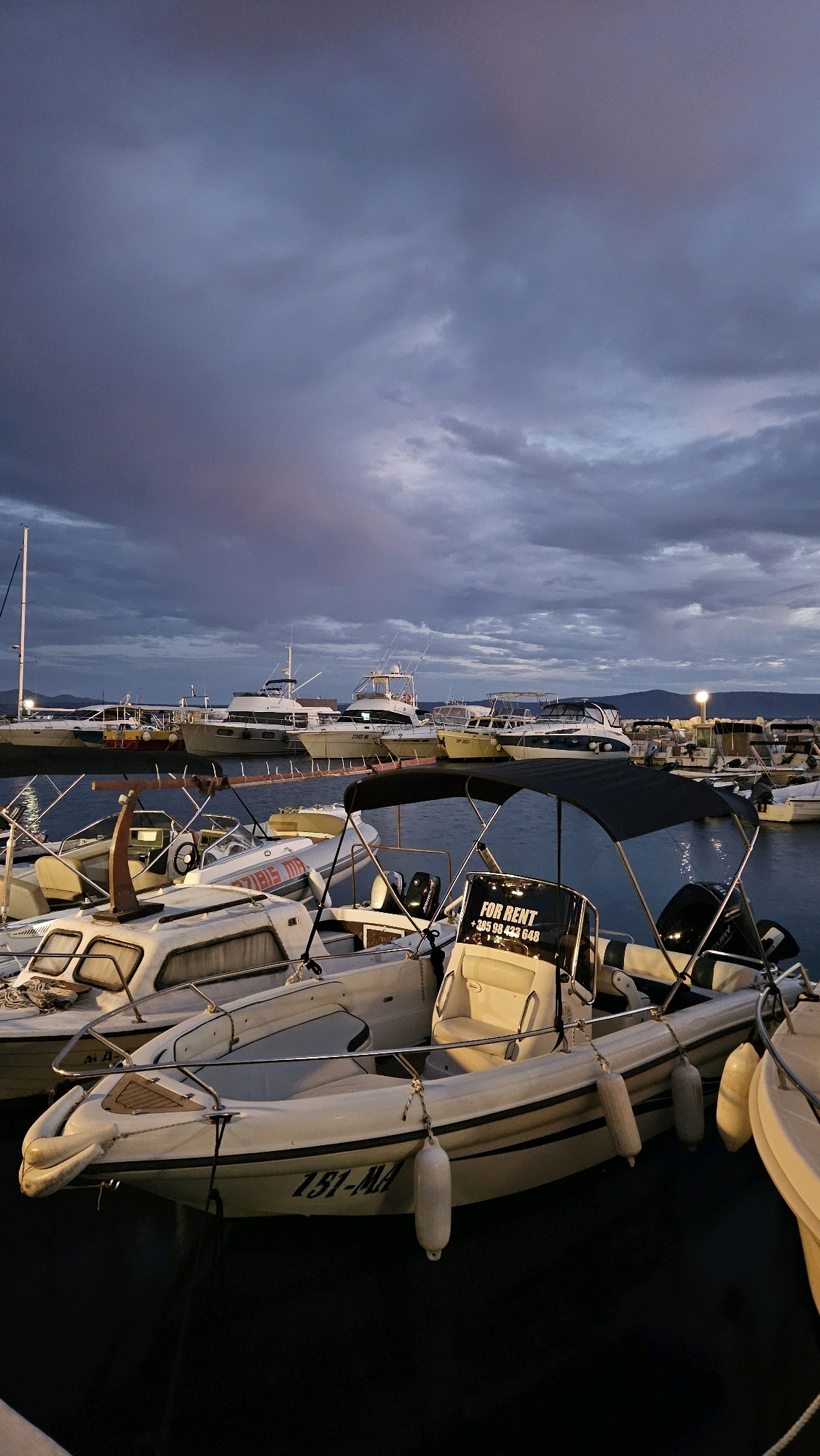 The image captures a harbor scene at dusk in Općina Tučepi, Croatia. In the foreground, several white motorboats are docked. One boat prominently displays "151-MA" on its hull and has a black canopy. Another boat behind it shows "FOR RENT" and a phone number on its windshield. Further back, larger yachts and smaller boats are moored in rows, silhouetted against the darkening sky. The sky is filled with dramatic, cloudy formations, with hints of pink and purple light suggesting the last rays of sunset. A tall mast of a sailboat is visible on the left. The overall atmosphere is calm, with the boats resting on the dark water.