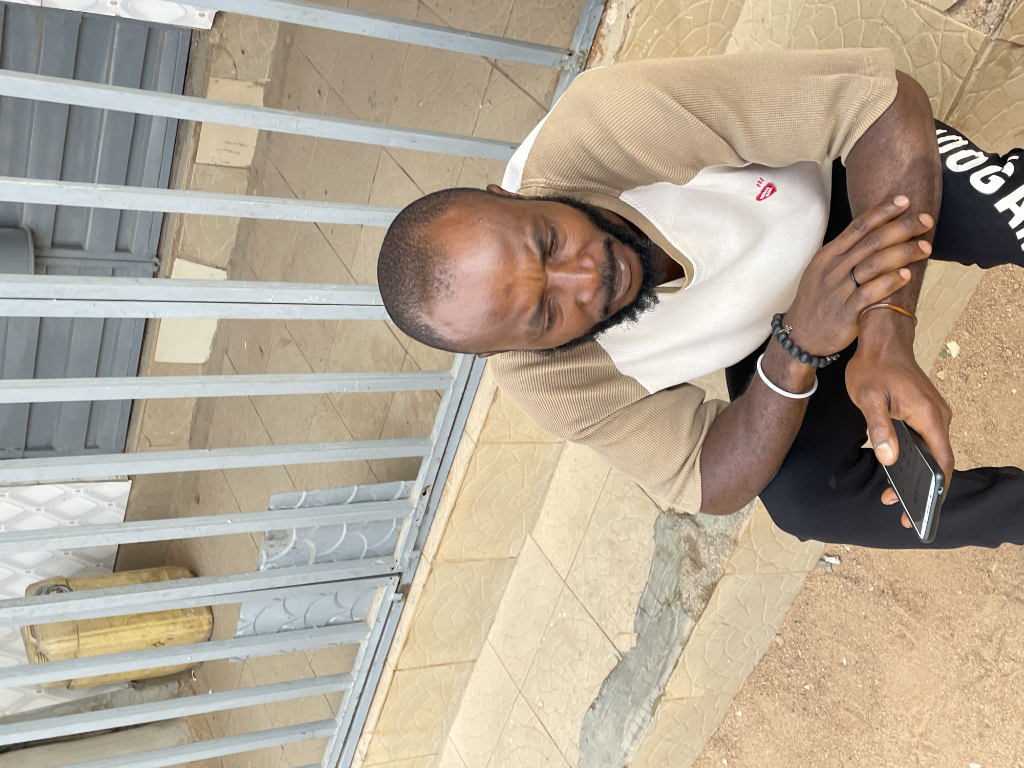 A medium shot shows a man sitting on outdoor tiled stairs. He is Black, bearded, and has a shaved head. The man is wearing a beige and white long-sleeved shirt, black sweatpants with white lettering, and multiple bracelets. He holds a black mobile phone in his right hand. Beside him are a gray metal fence and a yellow canister. He is located in Jos, Nigeria.