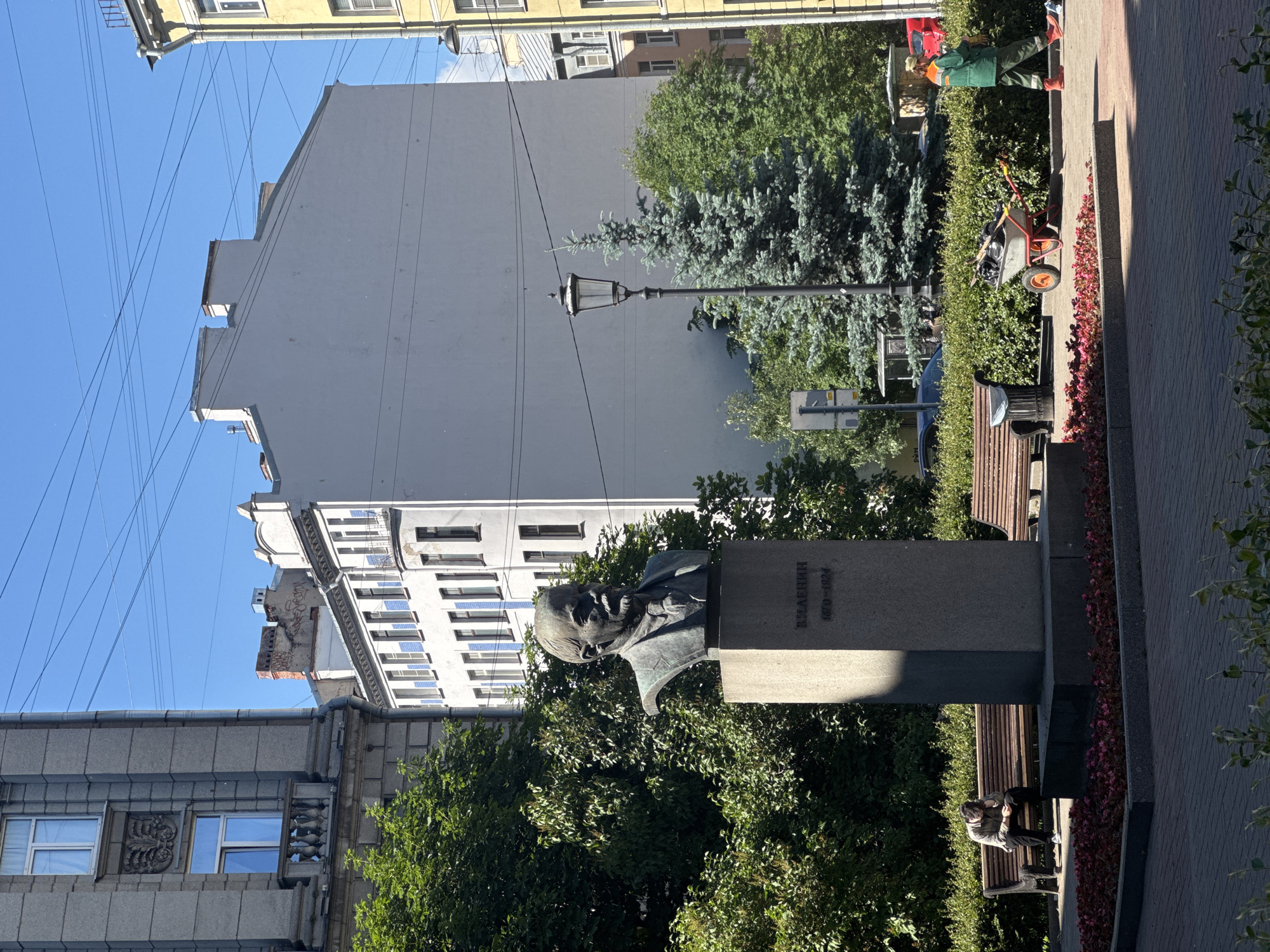 The image depicts an outdoor scene in an urban environment. In the foreground, a bust of a man with a beard and tie stands on a stone pedestal inscribed with Cyrillic text and dates. To the left of the pedestal, a person sits hunched over on a wooden park bench, their head resting on their hands. Further to the left, a large green tree provides shade. Behind the bust, a tall, slender lamppost stands beside a blue-green coniferous tree. In the middle ground, a low, trimmed green hedge lines the area. A worker in a green uniform and orange boots pushes a wheelbarrow laden with debris. Another park bench is visible to the right of the hedge. Several buildings of varying architectural styles and heights are visible in the background under a clear blue sky. One building on the far left is tall and grey with multiple windows. Adjacent to it is a white building with numerous windows and some blue decorative elements. Further back, a lighter gray building has a simple, flat roofline. A yellow building with windows is partially visible on the far right. Power lines crisscross the sky.