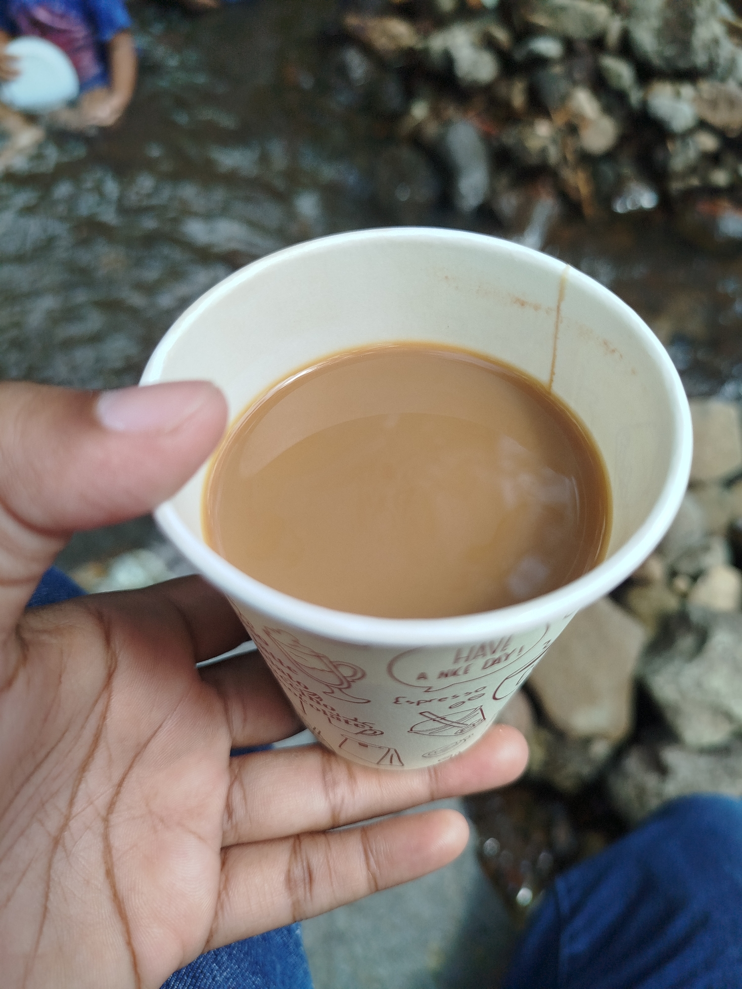 A close-up shot captures a hand holding a disposable paper cup filled with a light brown liquid, presumably tea or coffee. The cup has a pattern of small illustrations and text, including "HAVE A NICE DAY!". The background is out of focus, but it appears to be an outdoor setting with a shallow body of water and rocks. In the upper left corner, a person's legs and torso are visible, with one person wearing a blue shirt and another person holding a white object. The scene appears to be near a stream or river in Ngesrepbalong, Indonesia.