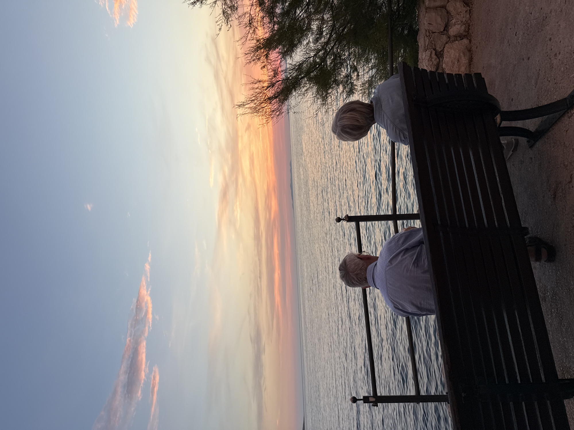 Two elderly individuals are seated on a dark wooden bench facing a body of water, presumably the Adriatic Sea. The man on the left, with gray hair and glasses, wears a light blue collared shirt and dark sandals. The woman on the right has blonde hair and wears a gray top. They are positioned with their backs to the camera, gazing at a sunset.
The sky displays a gradient of colors, transitioning from pale blue at the top to hues of pink, orange, and yellow near the horizon, indicative of a sunset. Wispy clouds are visible in the sky, catching the warm light. The sea is calm, with gentle ripples reflecting the colors of the sky. A dark metal railing runs along the edge of the waterfront in front of the bench. To the right of the bench, a rocky embankment with a shrub is visible.
The bench itself is dark wood with a slatted design and is situated on a paved surface. The scene is set outdoors, likely in a coastal area of Primošten, Croatia, given the characteristic landscape and sea. The overall mood is peaceful and contemplative, with the couple seemingly enjoying the sunset together.
