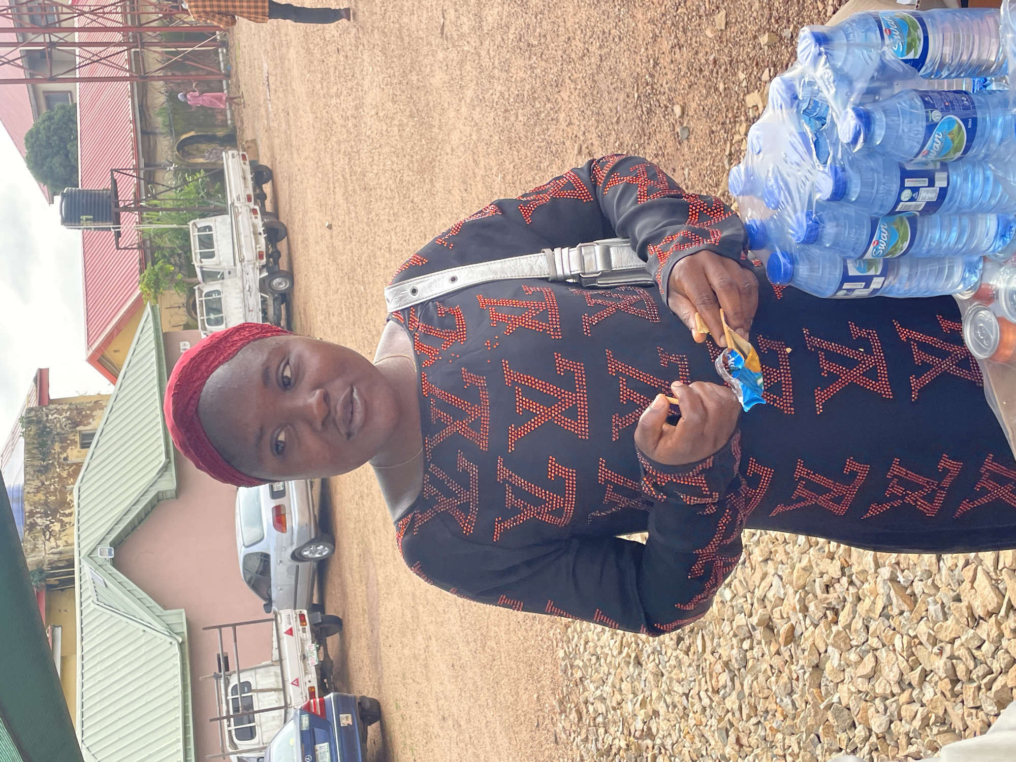 In Gura Top, Nigeria, a woman wearing a red headscarf and a black dress adorned with a red LV pattern stands in an outdoor setting. She holds a small object in her right hand, possibly a candy, and a silver bag is slung over her shoulder. In the foreground, a pack of Swan water bottles is visible. Behind her, several white trucks and cars are parked on a dirt and gravel surface. Buildings with red and green roofs are in the background, along with a water tower and a metal structure. The sky is overcast.